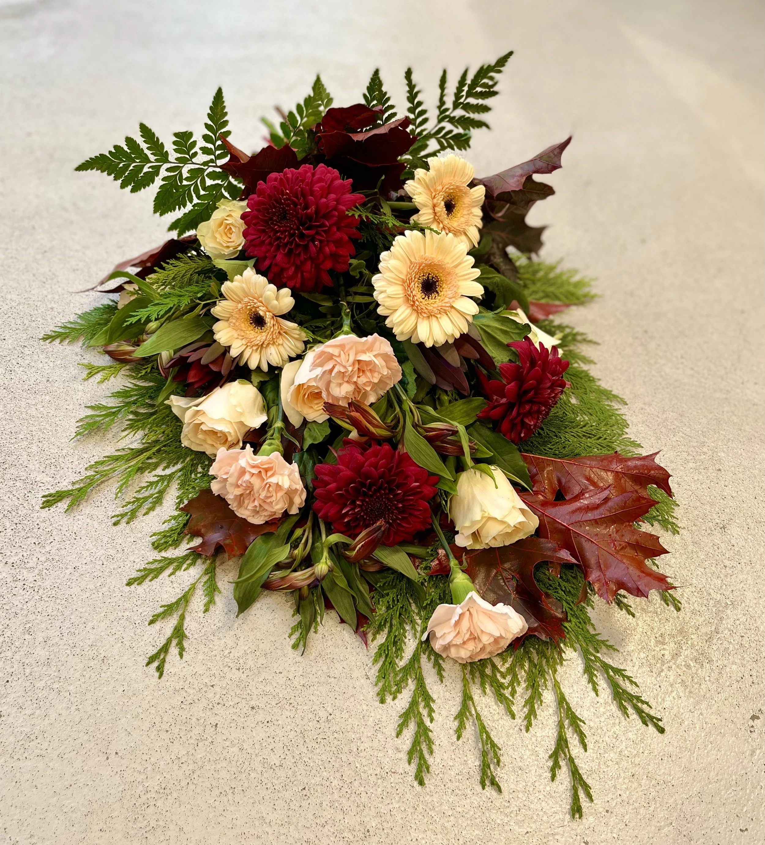 En bbegravningsbukett med röda, aprikosa och ljusrosa blommor samt gröna blad och barr.