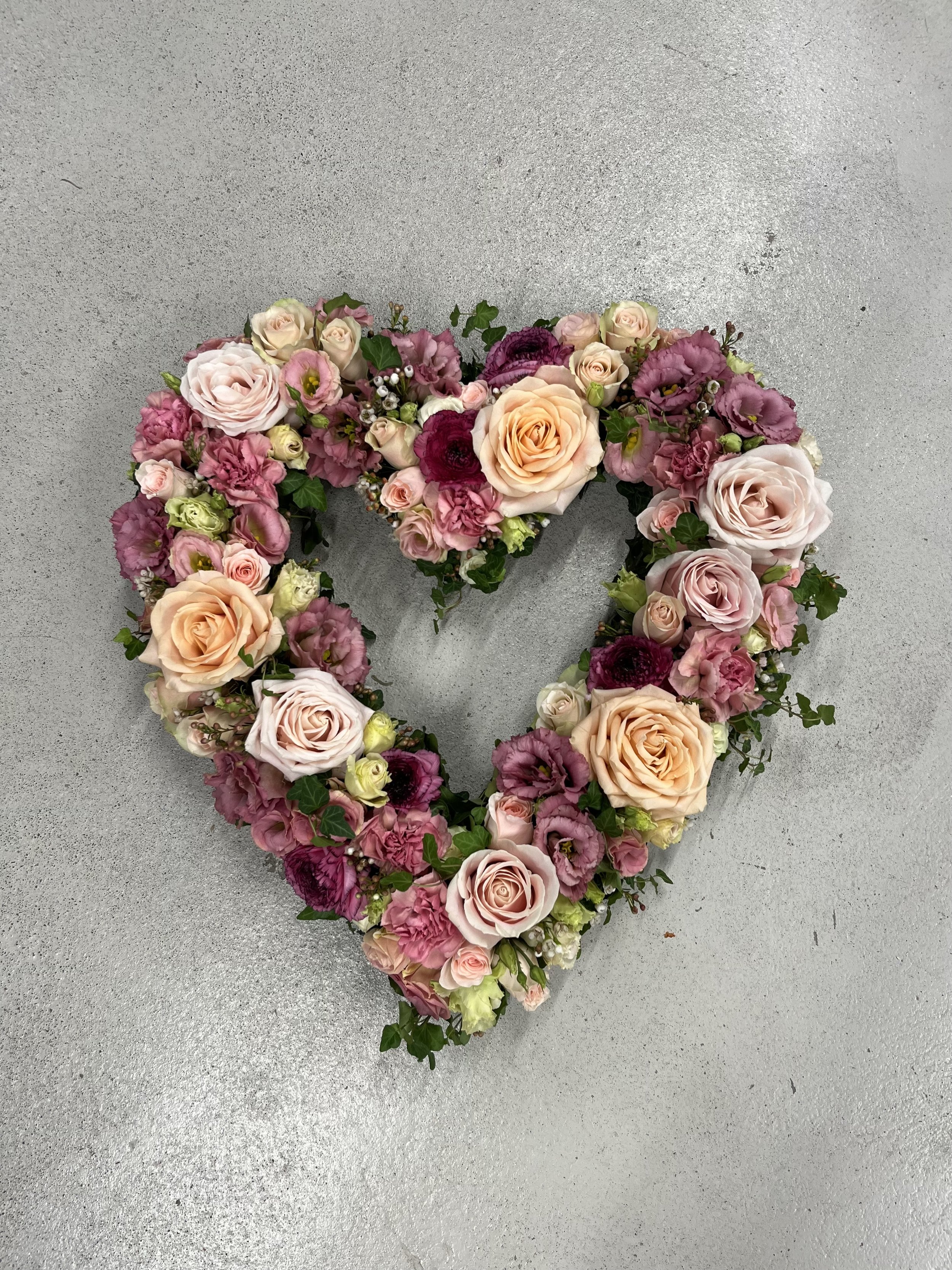 En blomsterdekorationi form av ett hjärta med rosa, vita och aprikosa blommor på en grå yta.
