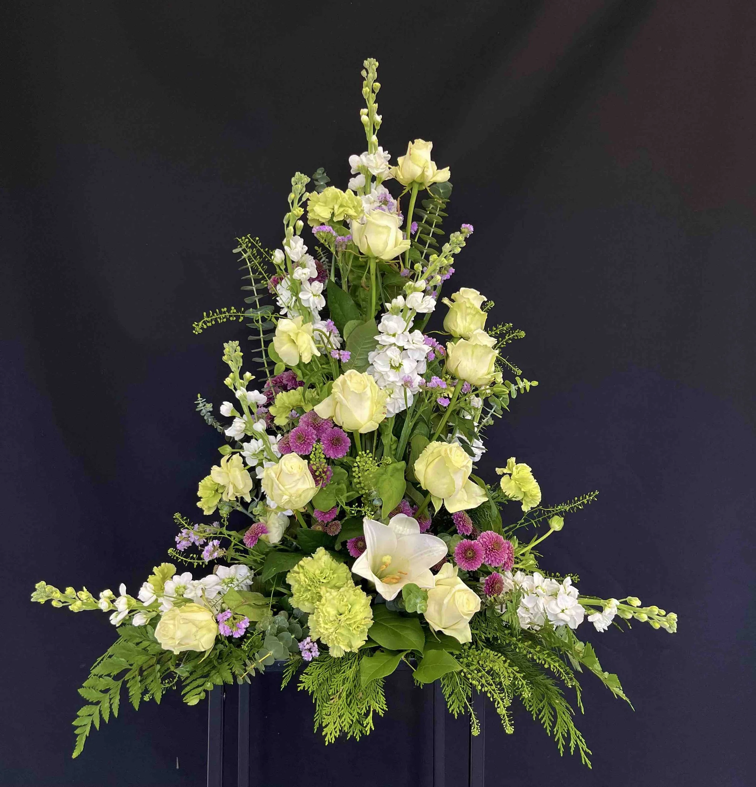 Hög begravningsdekoration med gul rosor, vita lövkojor, rosa och vita småblommor, och grönt bladverk mot en mörk bakgrund.