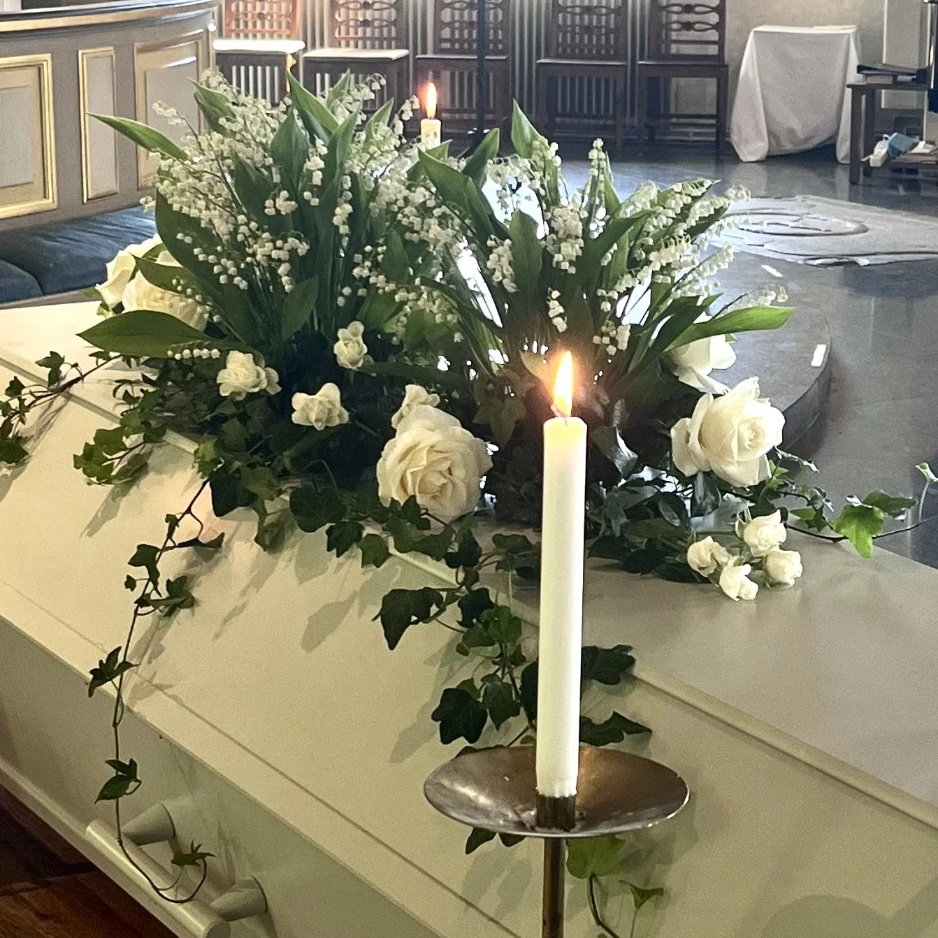 En vit blomsterarrangemang med vita rosor och liljekonvaljer på ett vitt skåp, med en brinnande ljus i förgrunden.