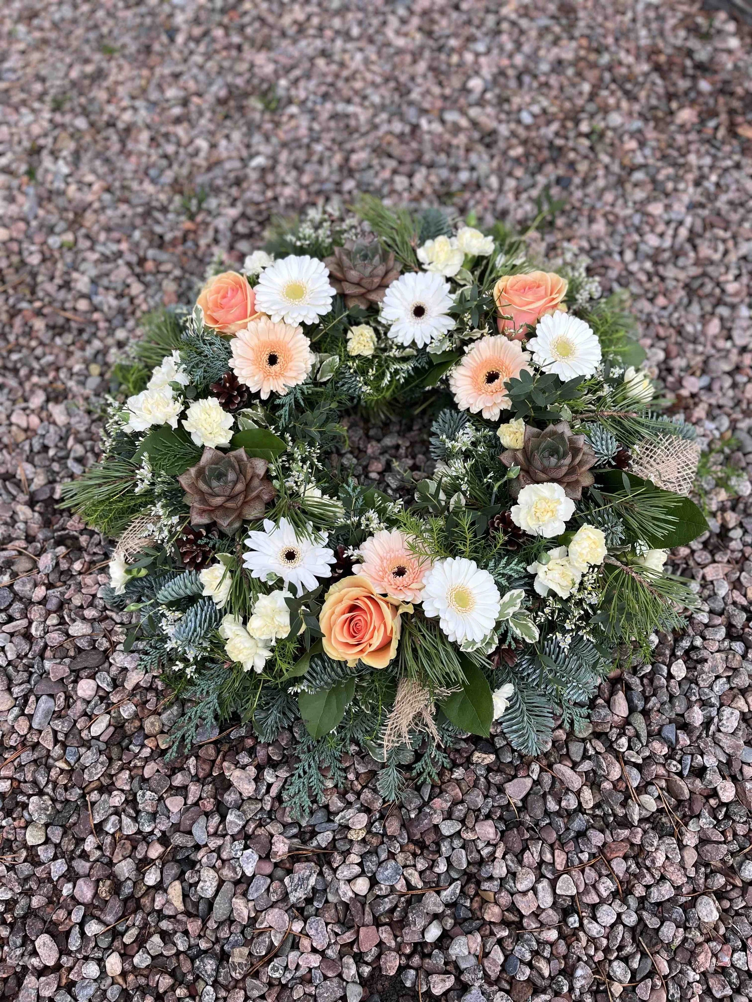 Krans som består av vita och aprikosa blommor samt grönt löv och barr.