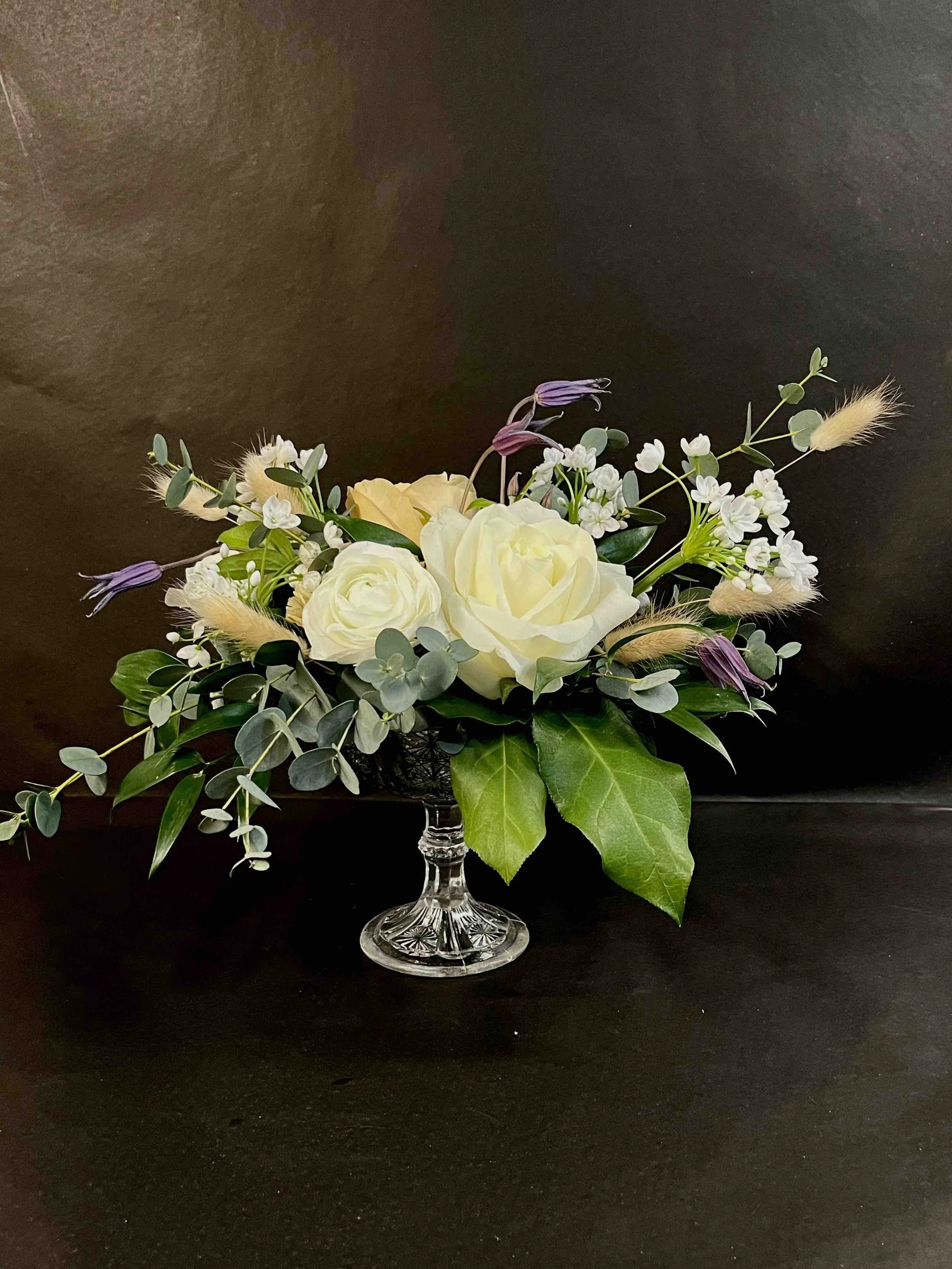 En blomsterarrangemang i en kristallvas, bestående av vita rosor, gröna blad, eucalyptus och små vita blommor, på en svart bakgrund.