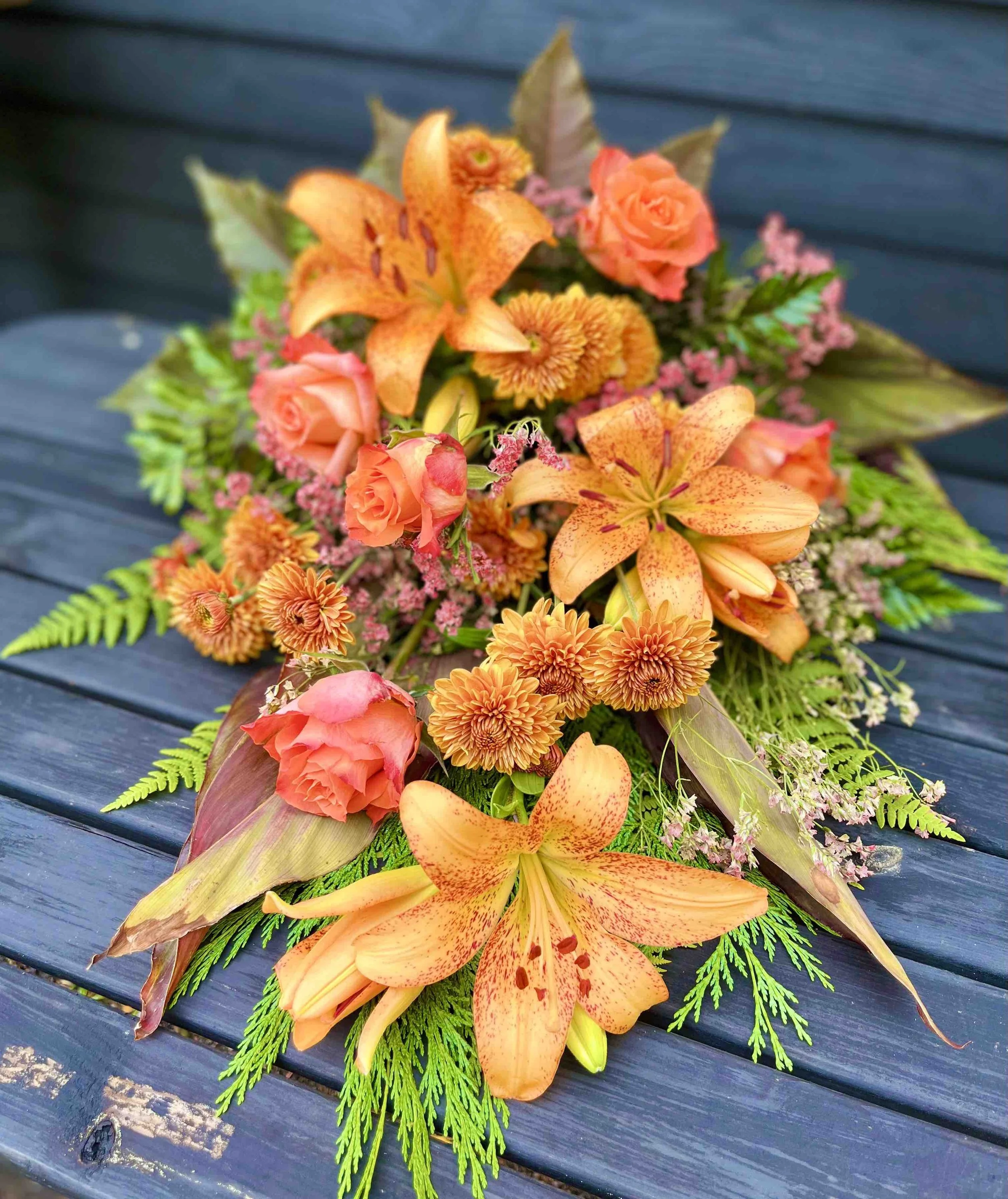 En höstig liggande bukett för begravning med orange liljor, rosor och chrysanthemum på en mörkblå träbänk.