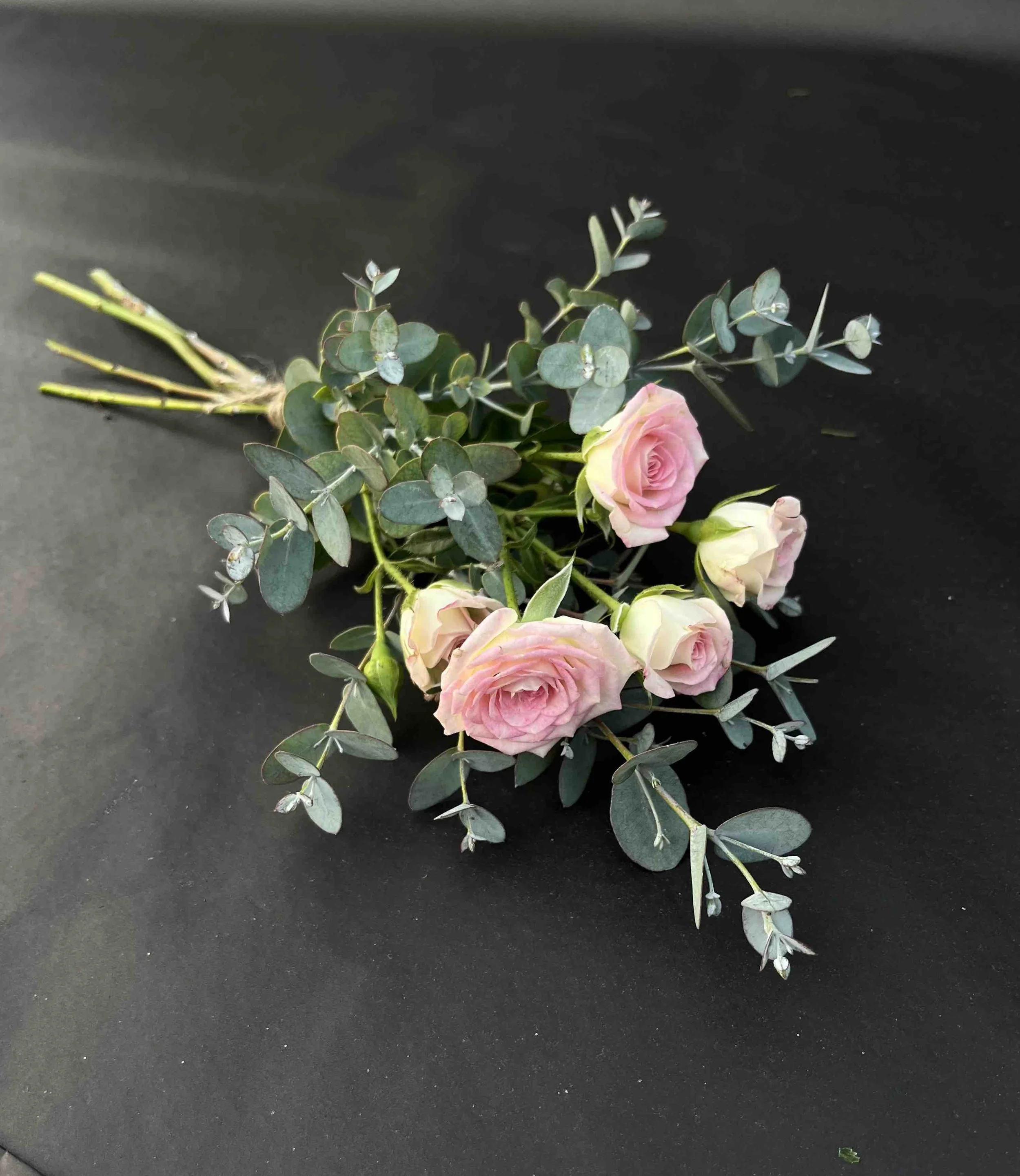 En liten handbukett för begravning med ljusrosa kvistrosor och grön eukalyptus på en svart bakgrund.