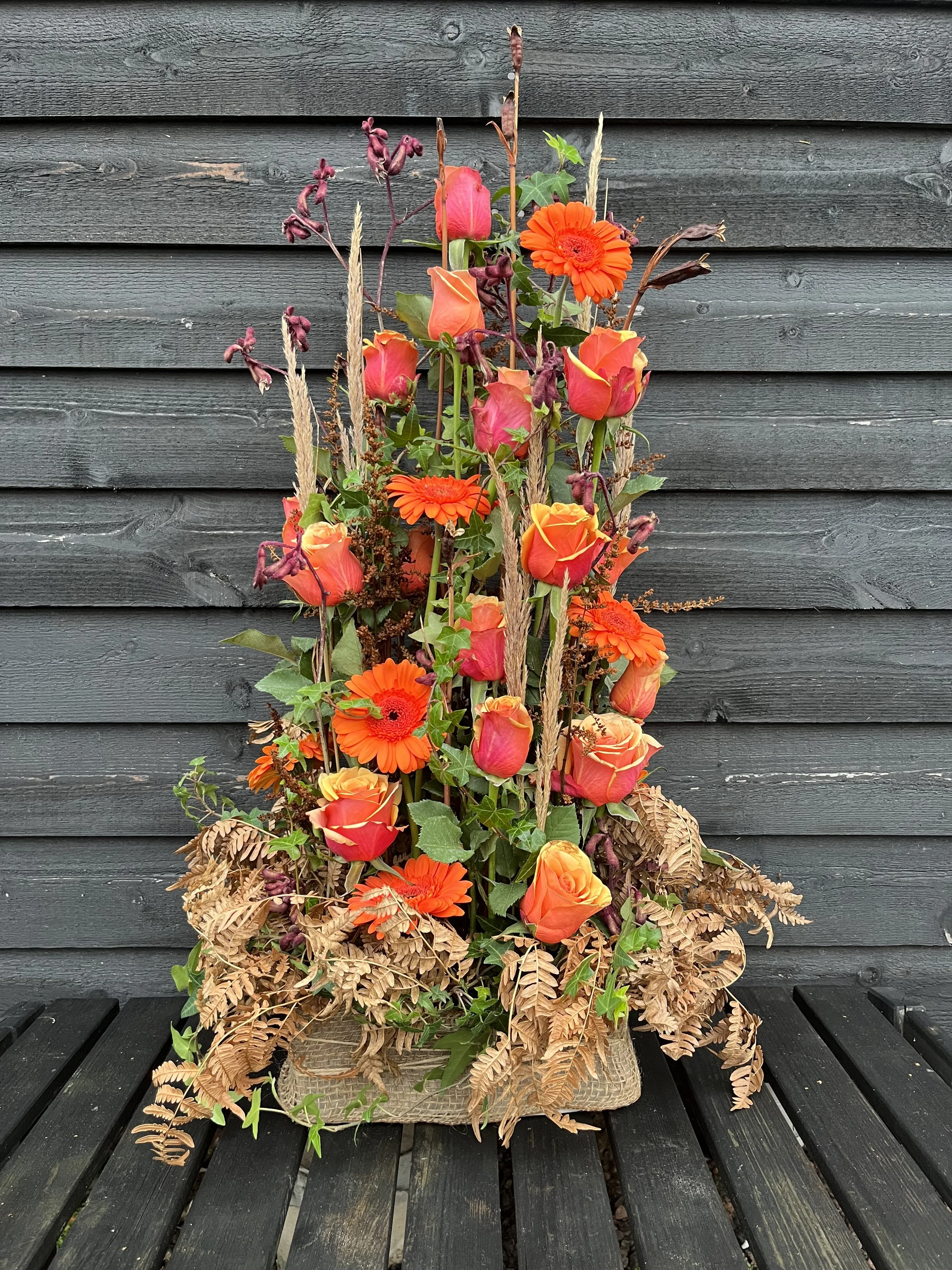 En parallel, hög dekoration för begravning med orange rosor och gerbera på en svart träbänk framför en mörk trävägg.