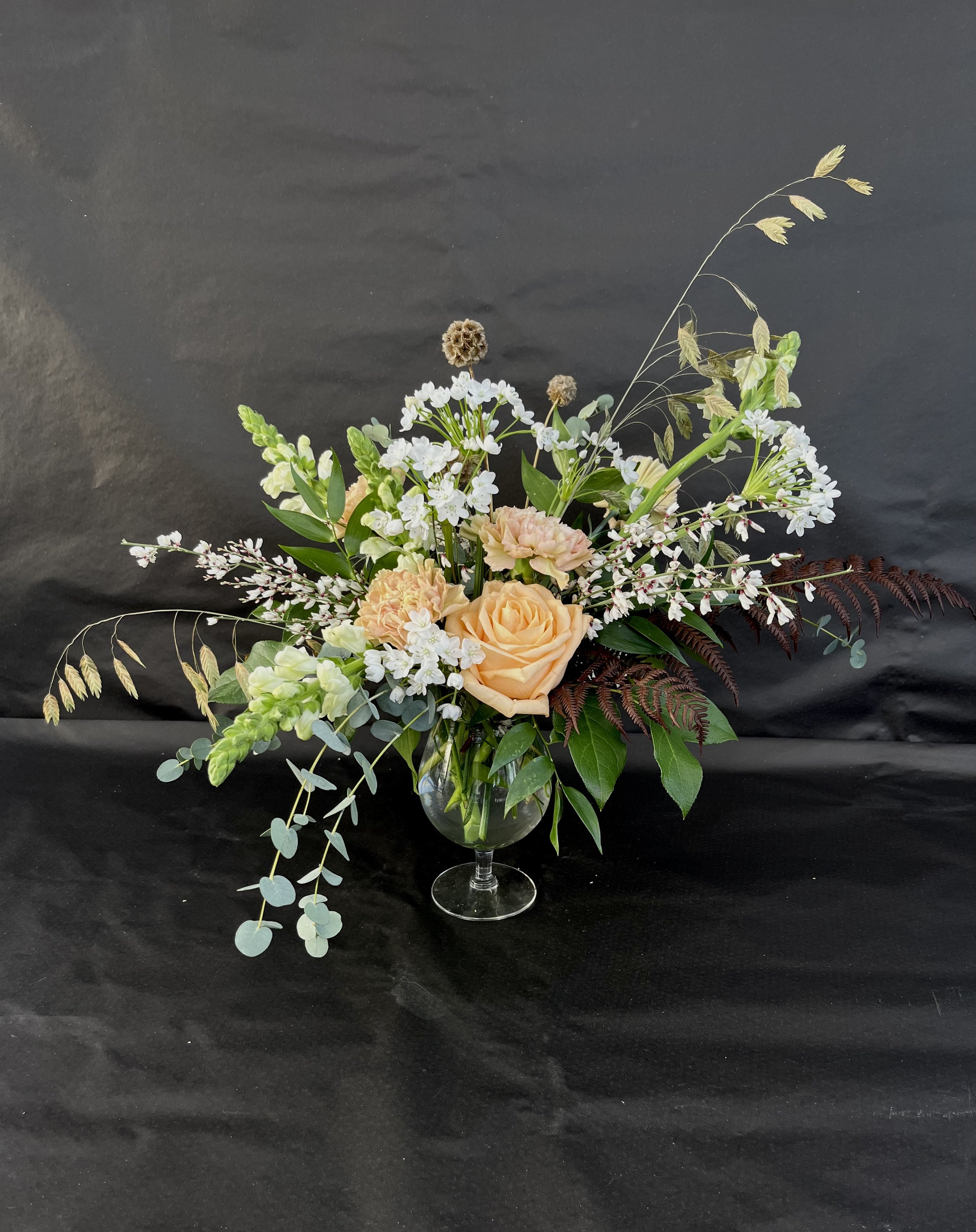 Blommig vas med beige och vita blommor, gröna blad och torkade växter, på svart bakgrund.