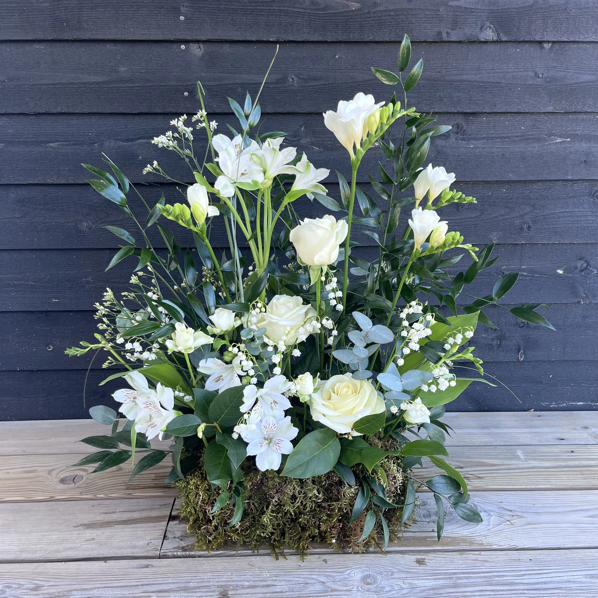 Blomsterdekoration med vita blommor och grönt löv i en mossig bas på ett träbord framför en svart trävägg.