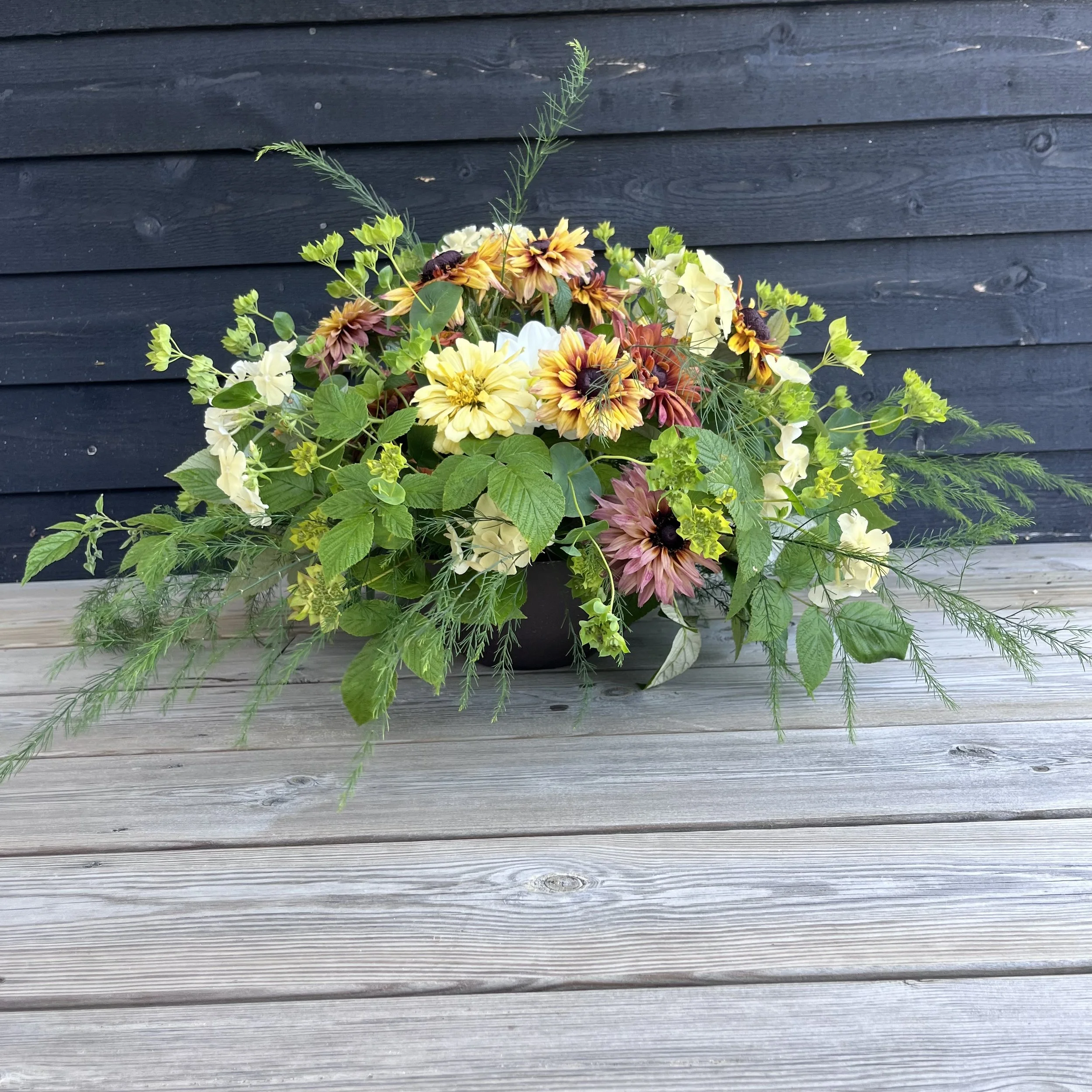 Färgglad blomsterarrangemang i en svart kruka på en träbänk med en mörk vägg i bakgrunden.