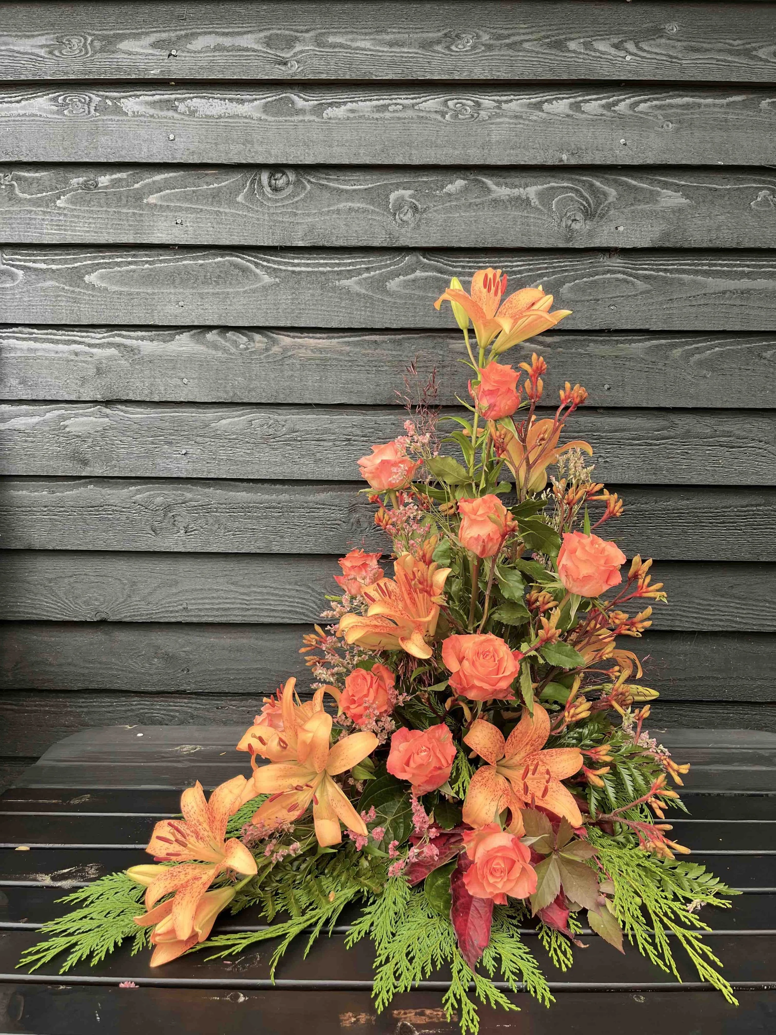 En asymetrisk begravningsdekoration med orange liljor och rosor.