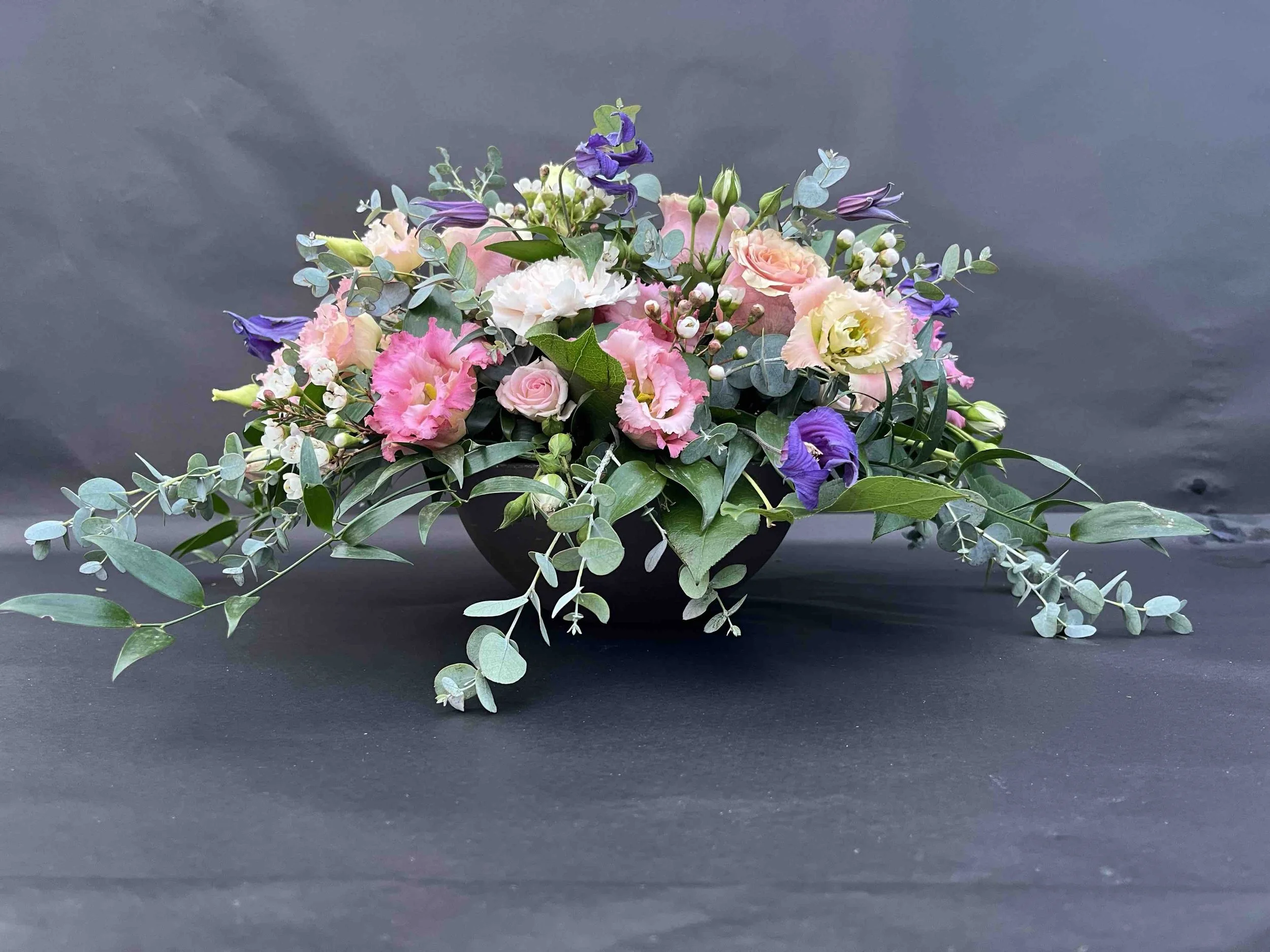 En miljövänlig begravningsdekoration i en bioskål med rosa, vit och lila blommor samt grönt bladverk.