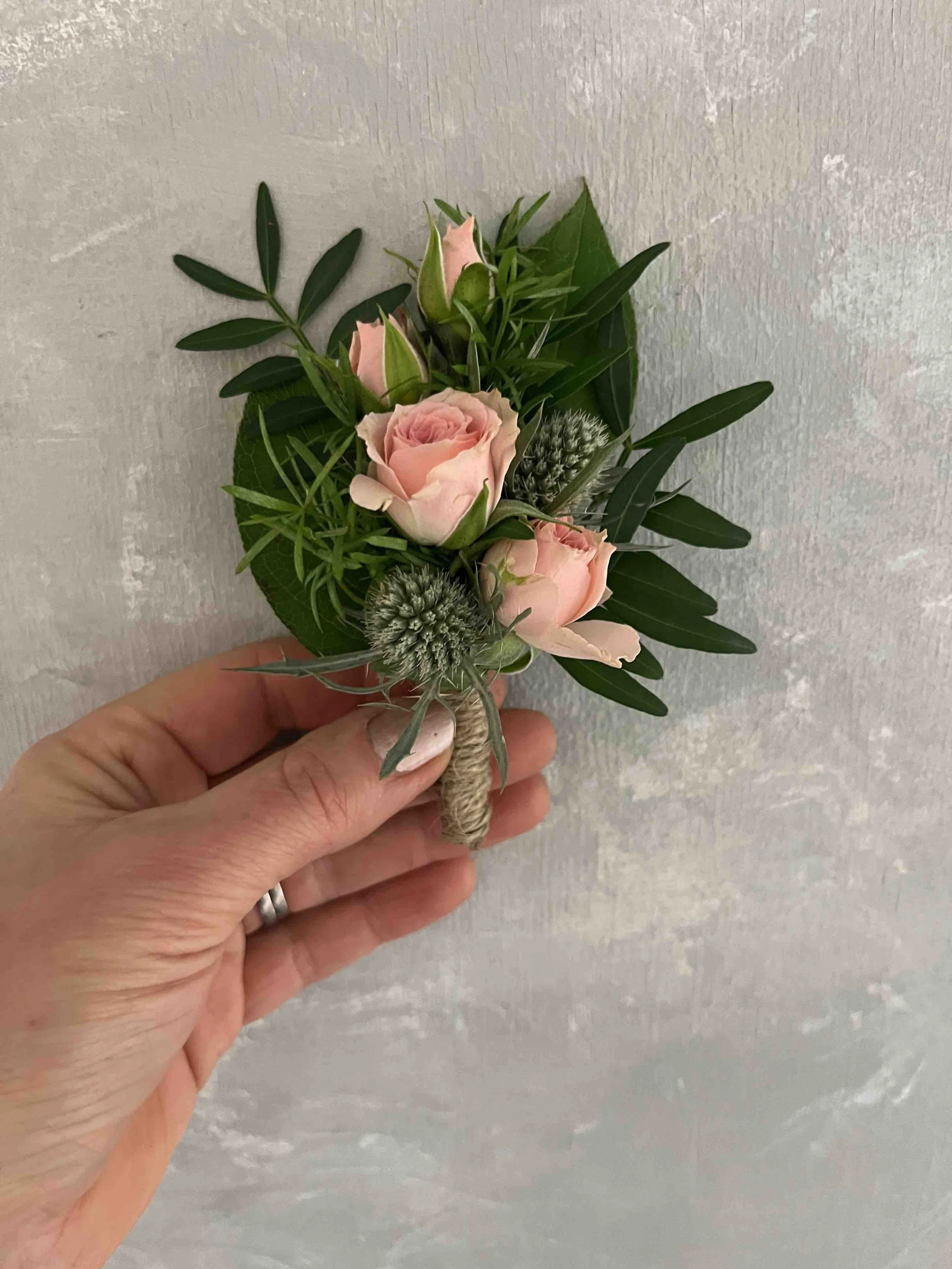 En hand håller ett rispaket med en liten bukett av ljusrosa rosor, gröna blad och nån form av pustel.