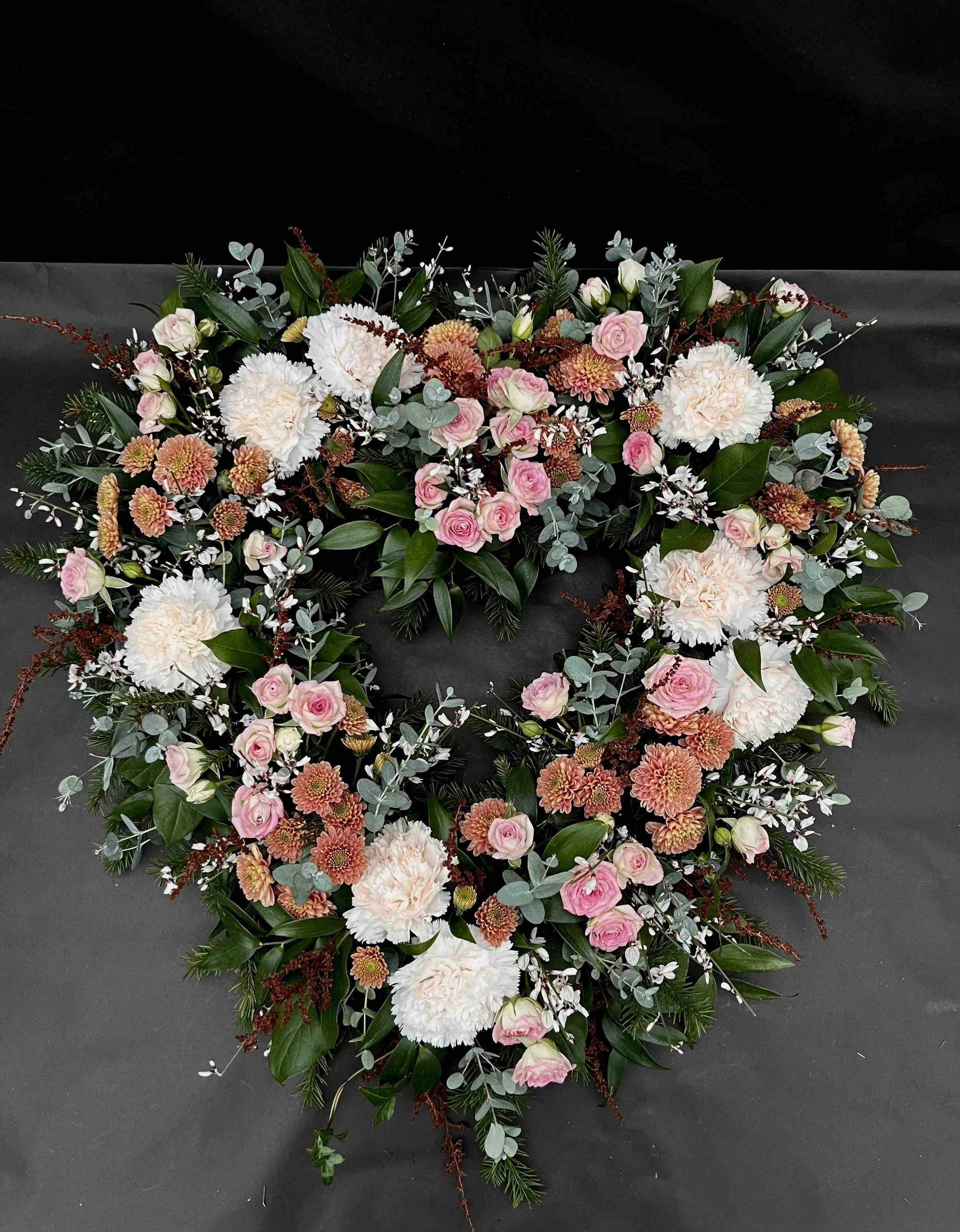 Blomsterkrans med vita och rosa blommor, gröna blad och silverfärgad bladverk mot svart bakgrund.