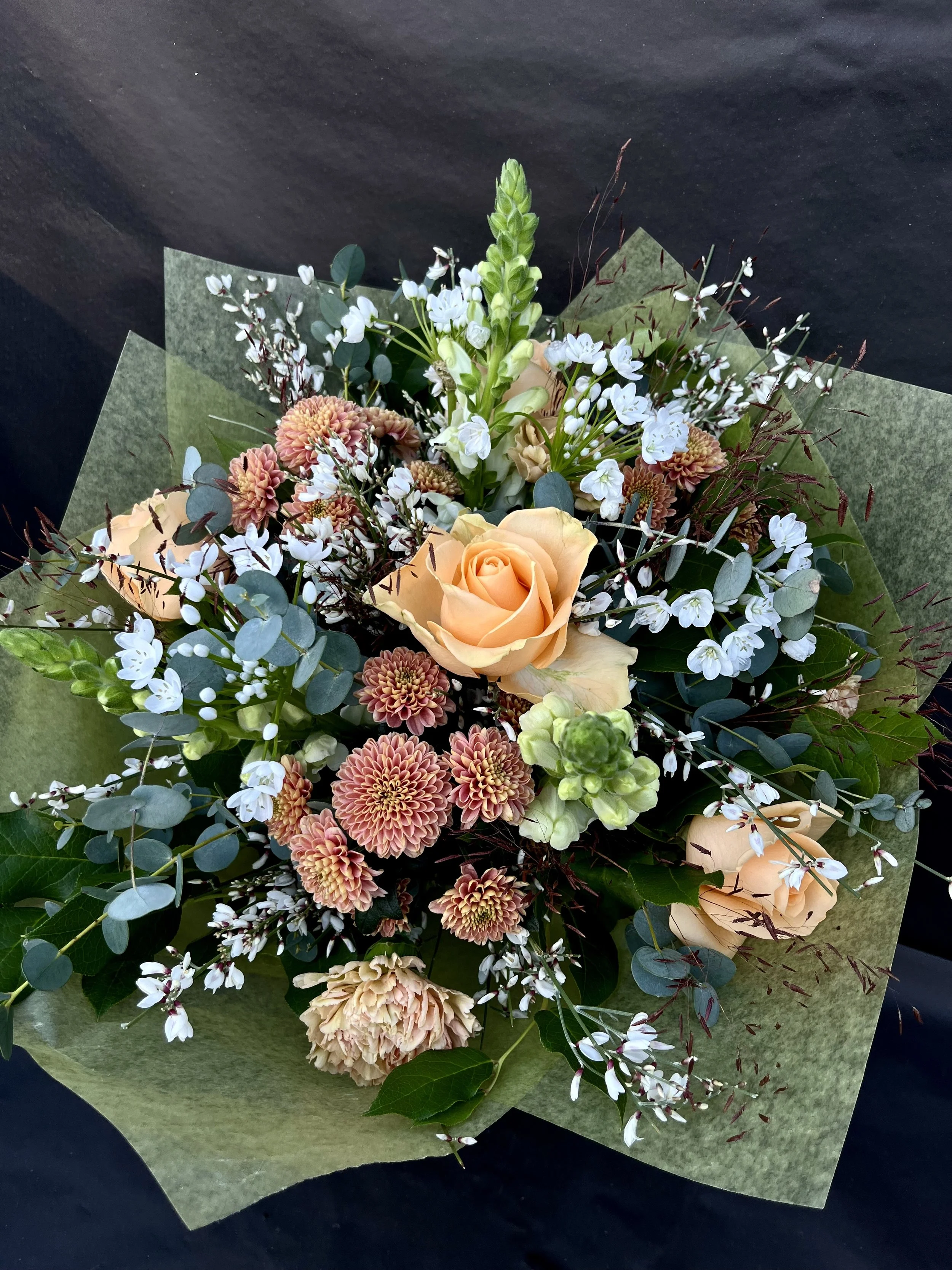 Blombud med aprikosa och vita blommor, inklusive rosor, chrysanthemum och vit ginst omgiven av gröna blad, presenterat i grönt silkespapper.