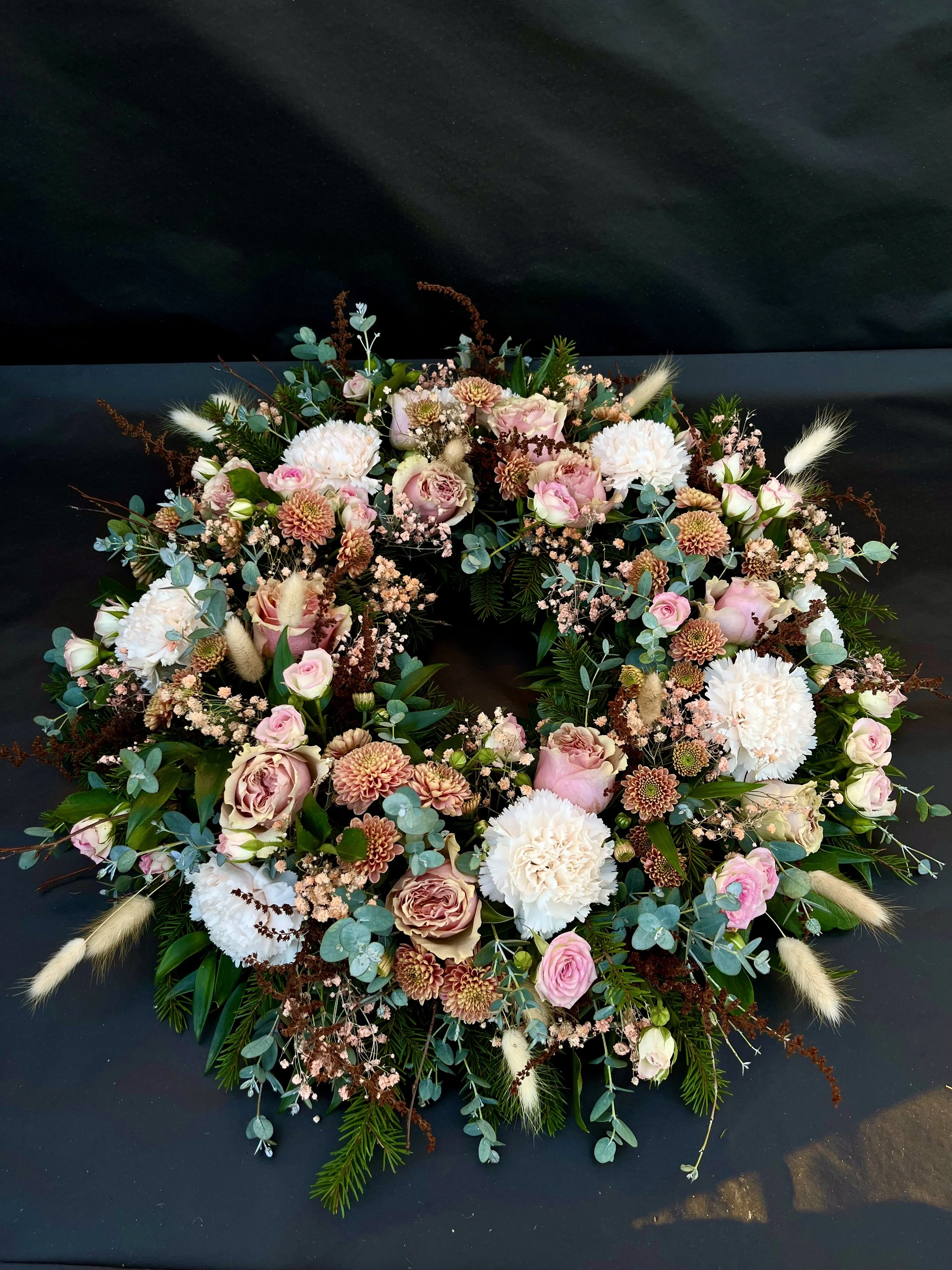 Blomsterkrans för begravning med rosa och vita blommor, gröna blad och gräs, mot en mörk bakgrund.