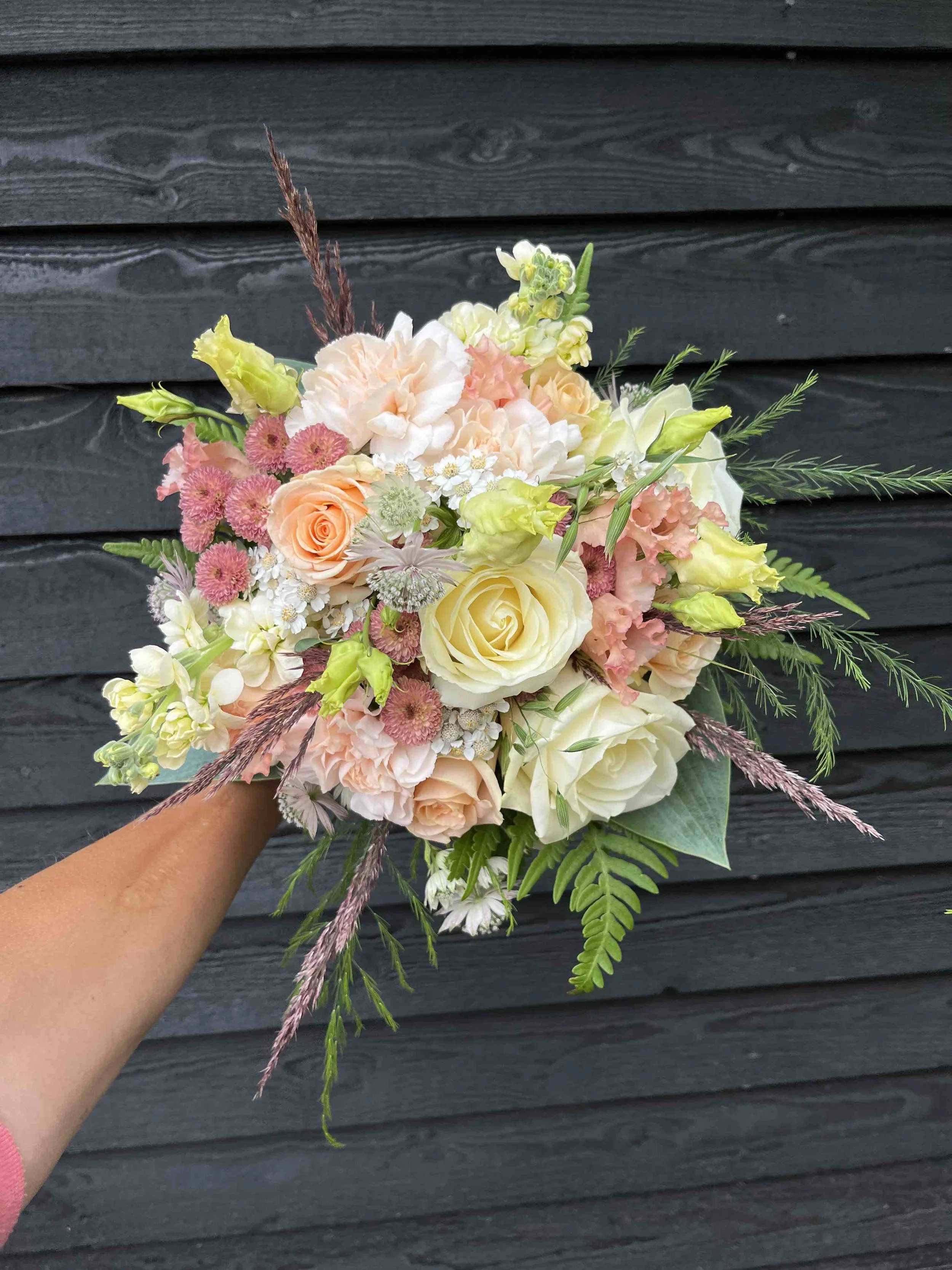 Hand som håller en bukett med vita, rosa och aprikos blommor framför en svart trävägg.