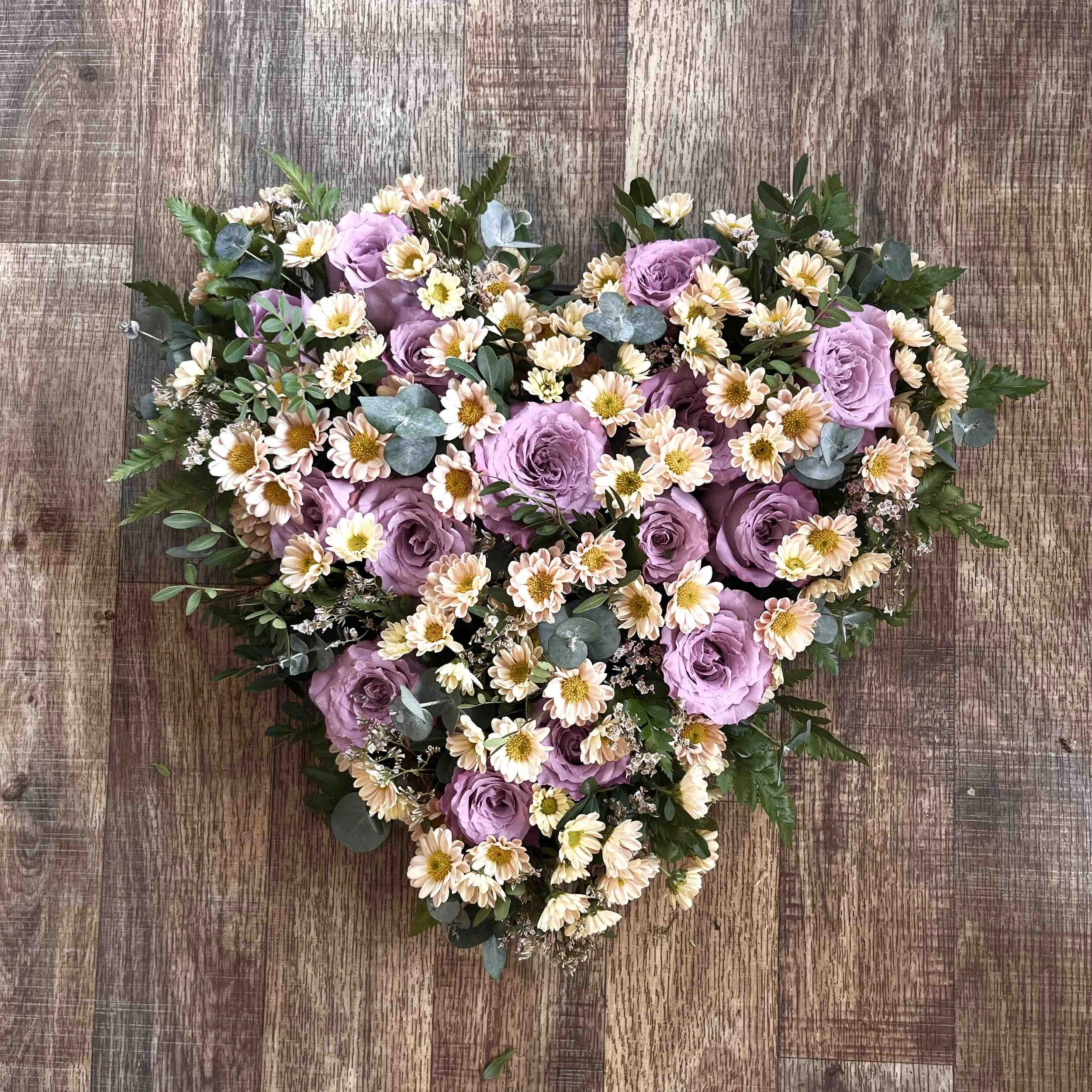 En hjärtformad begravningsdekoration med ljuslila och vita blommor med gröna blad, placerad på ett träbord.