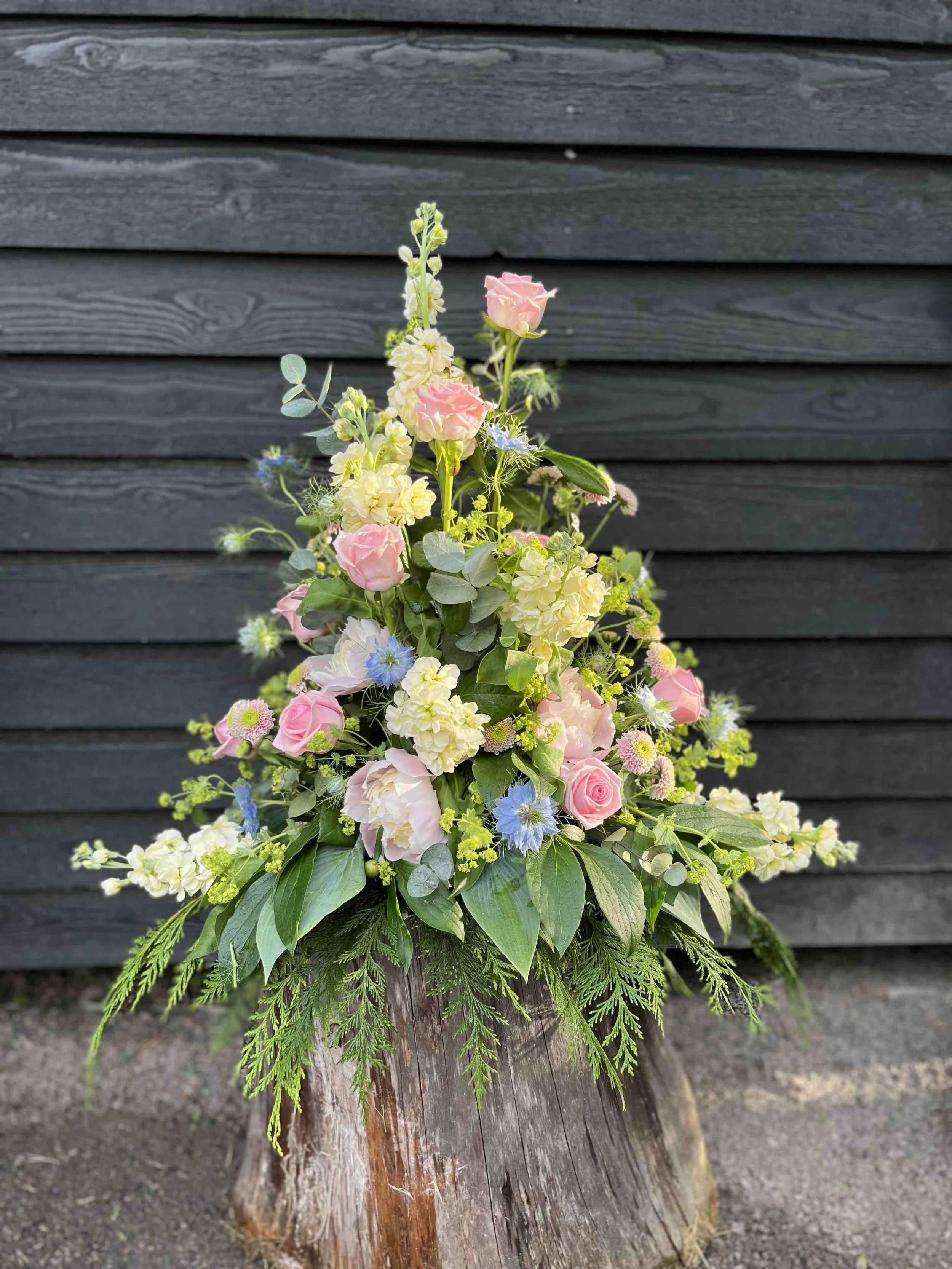 Hög vårig Begravningsdekoration med rosa, vita och blå blommor, placerad på ett trästycke mot en svart träpanel.