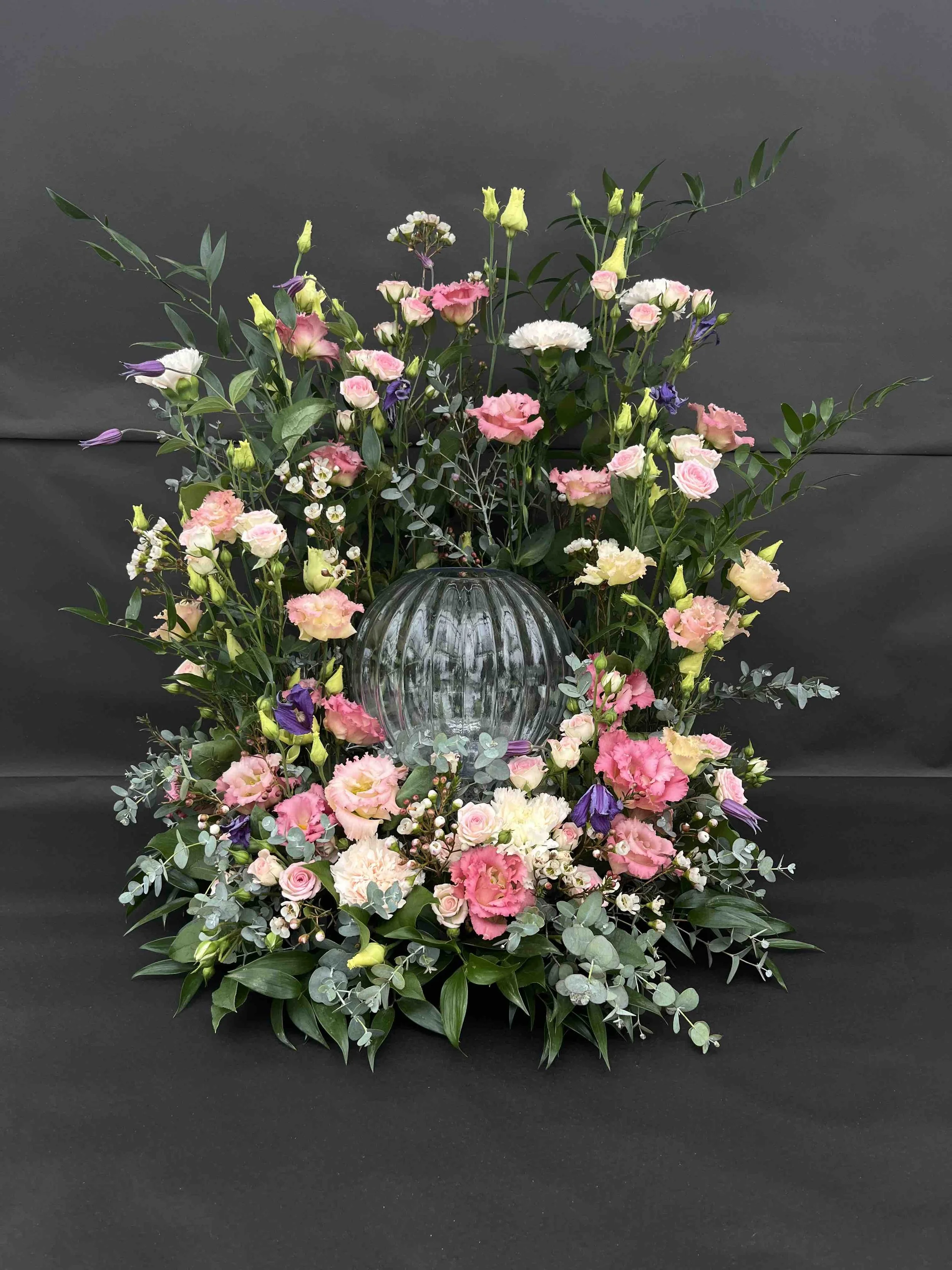 Urndekoration med ängskänsla bestående av rosa, vita, och lila blommor samt gröna blad.