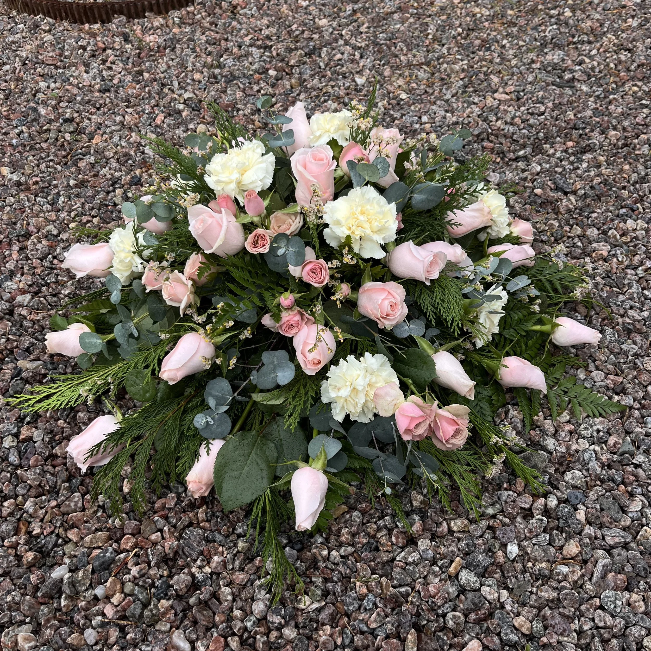 En  rund, låg begravningsdekoration med rosa rosor och creme vita nejlikor på grus.