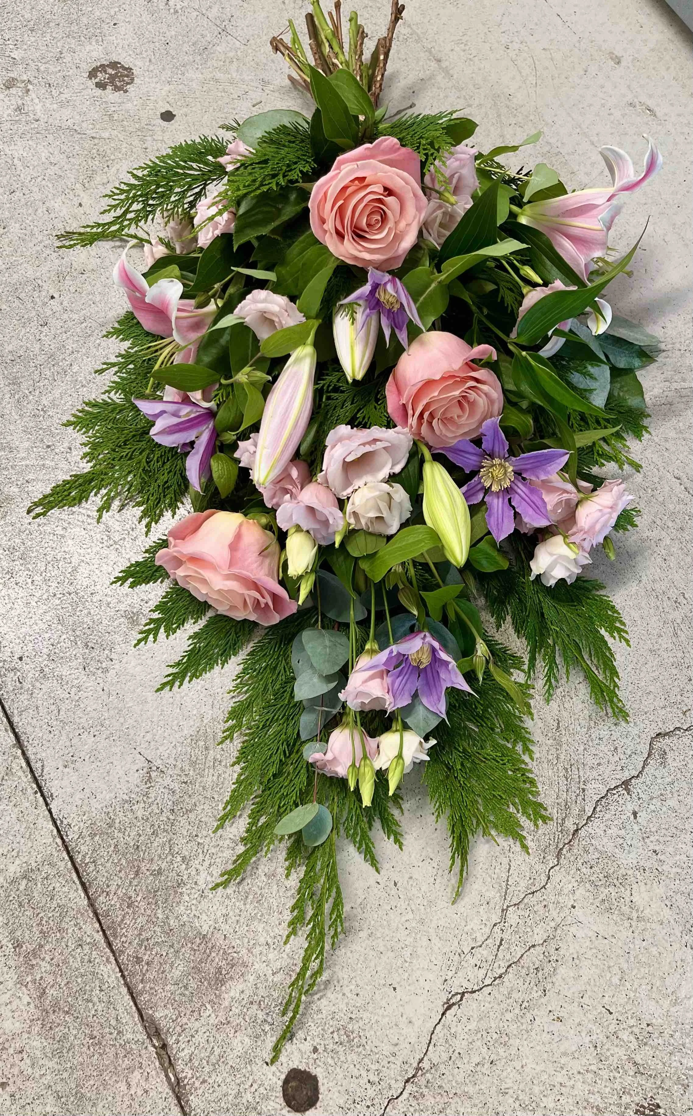 En bukett liggande med rosa rosor, liljor, lila clematis och gröna blad, placerad på en betongyta.