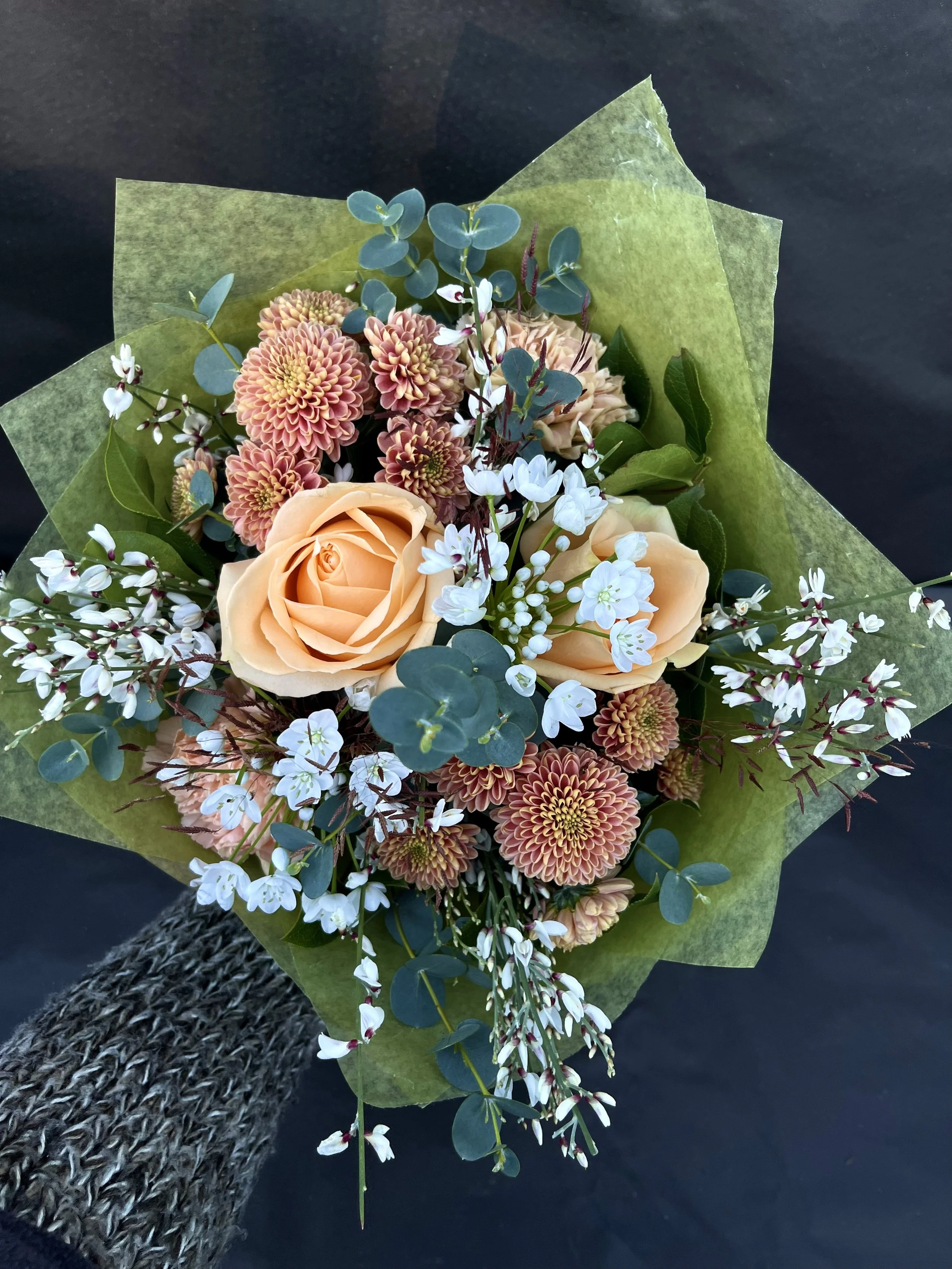 En bukett med ljusorange rosor, chrysanthemum, vit ginst och gröna blad, inbäddat i grönt blompapper.