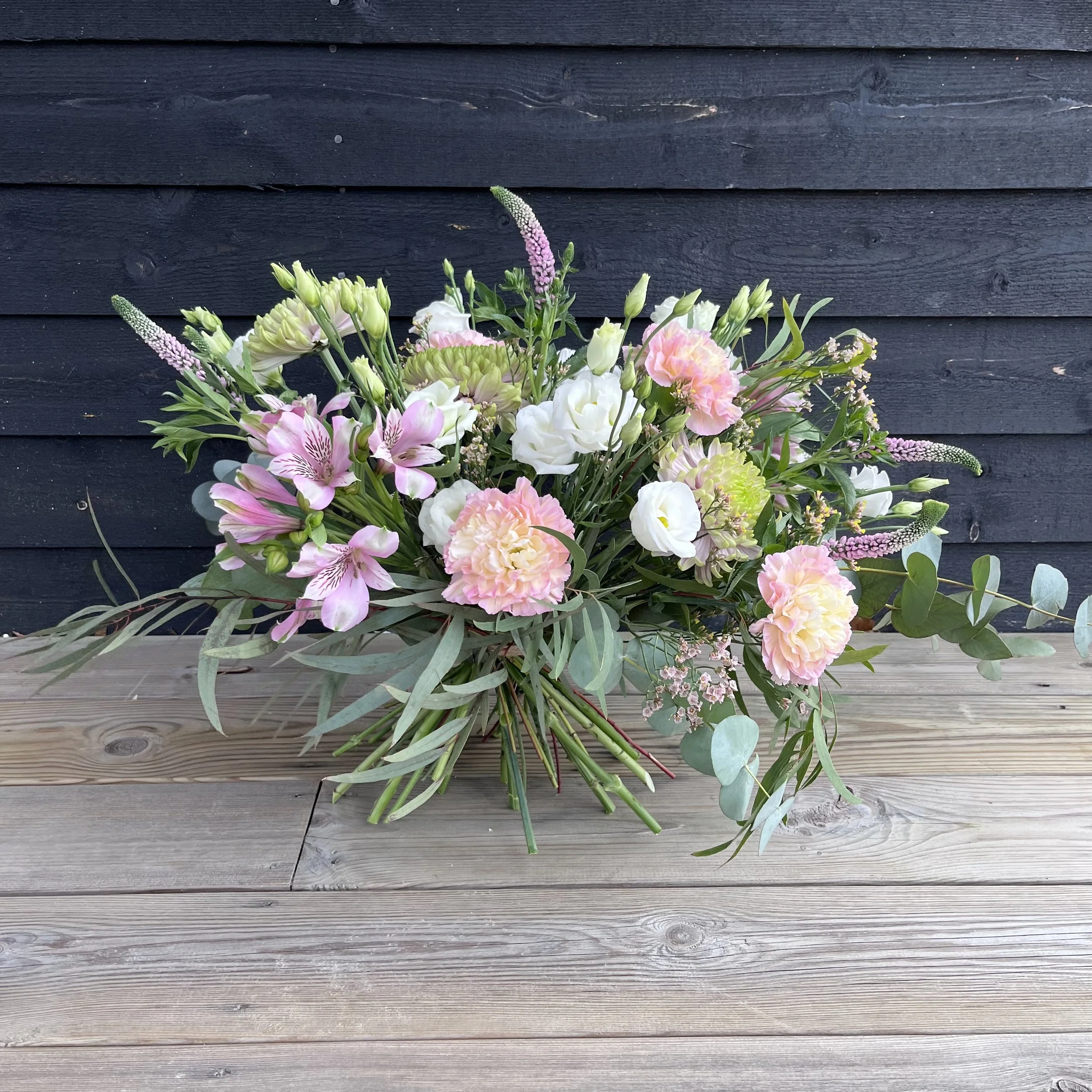 En bukett med rosa, vita och gröna blommor på ett träbord framför en mörk trävägg.