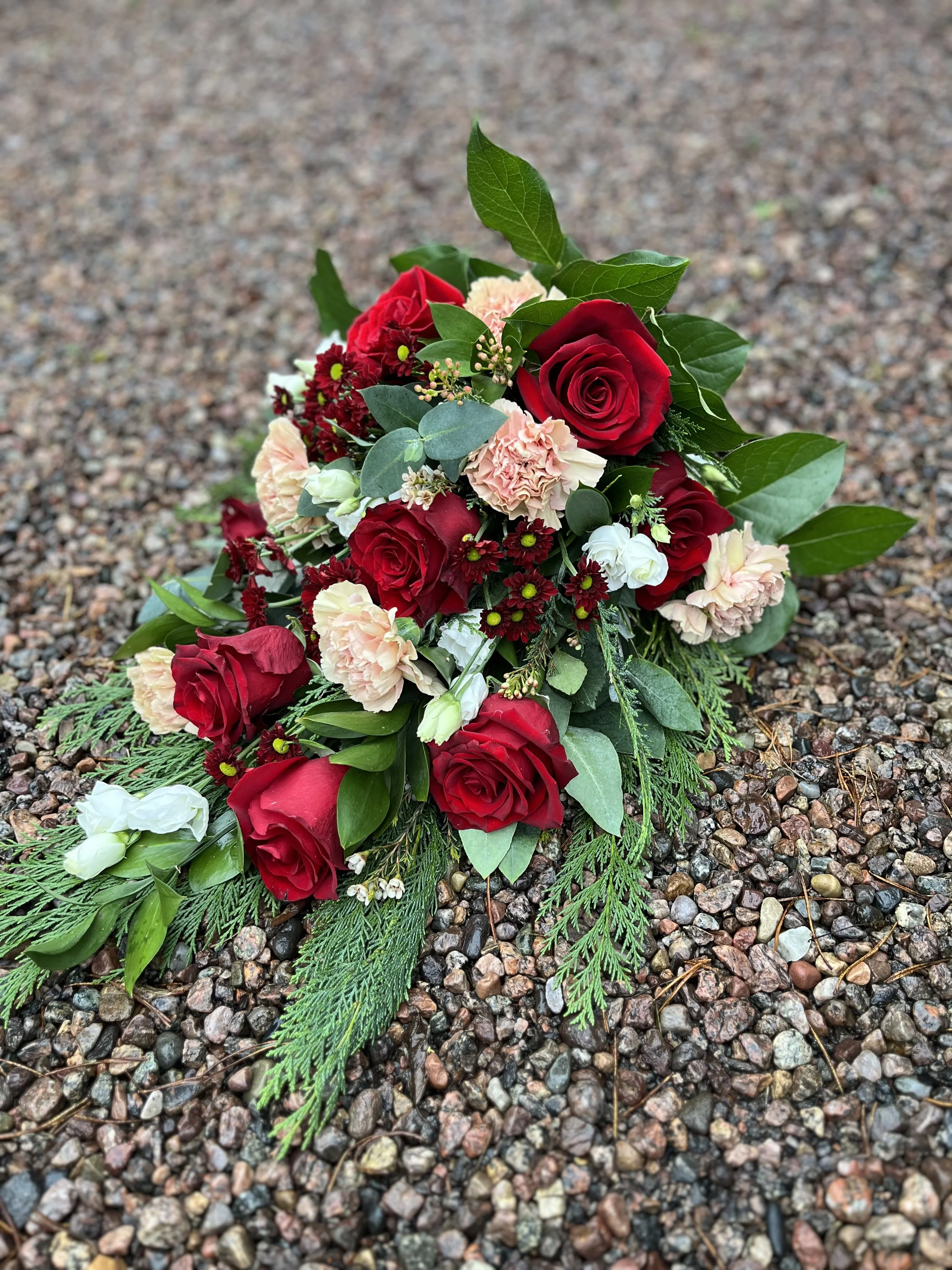 En liggande begravningsbukett med röda rosor, vita prärieklockor och rosa nejlikor.