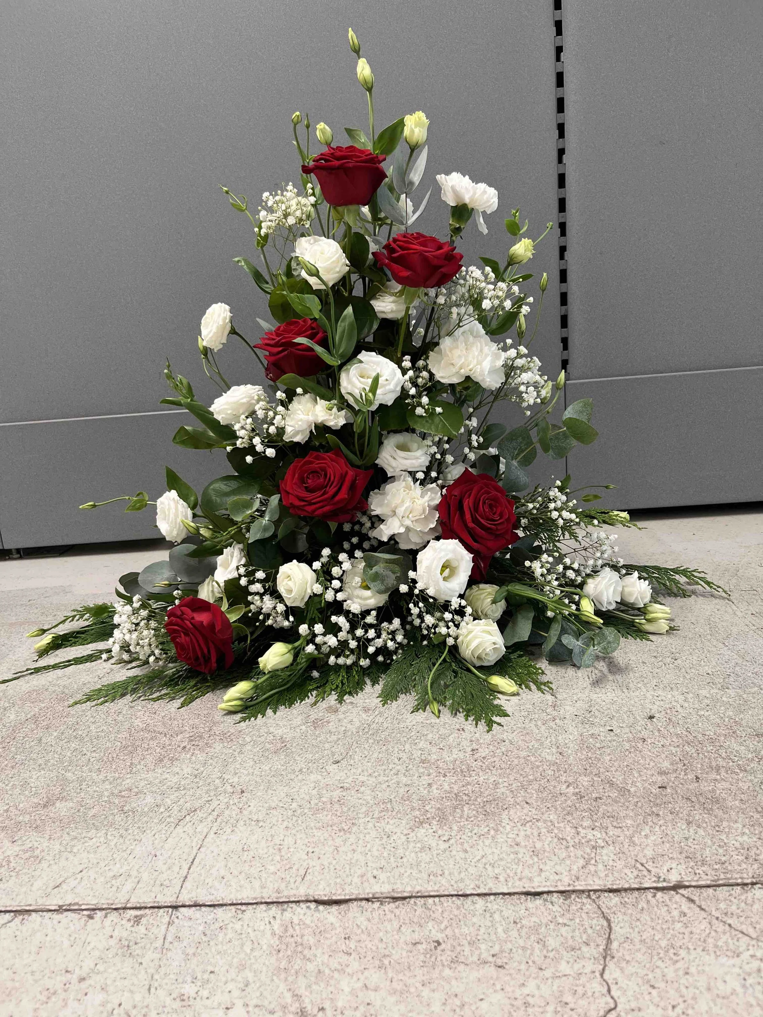 En blomsterdekoration för begravning med röda rosor, vita prärieklockor, baby’s breath och grönt blad, placerat på golvet mot en grå vägg.
