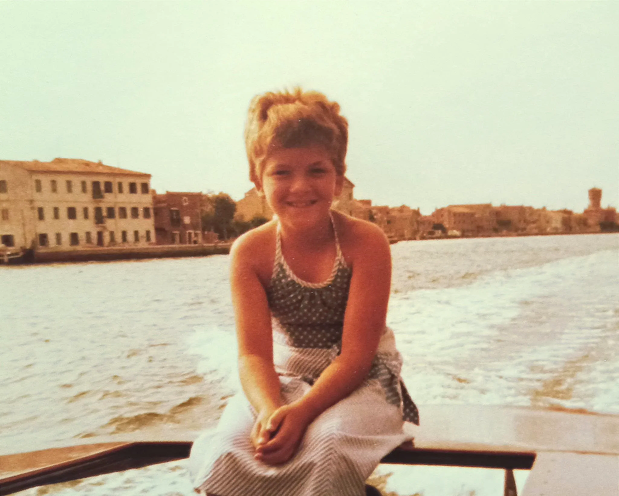 Een jonge meisje zit op een boot met water en gebouwen op de achtergrond, tijdens zonsondergang in de stad.