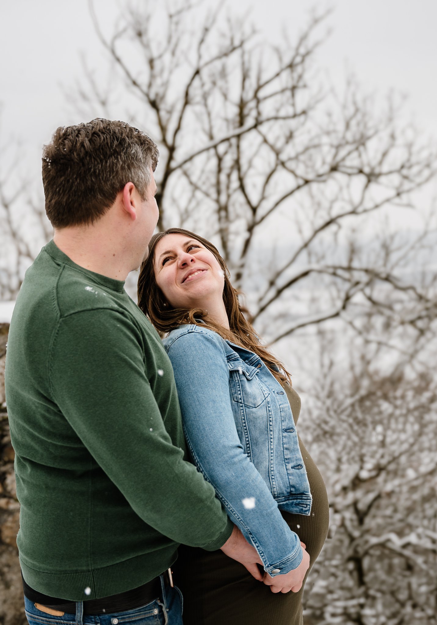 Ein Paar steht im Schnee vor einem winterlichen Baum, der keine Blätter hat, und sie schaut sich liebevoll an.