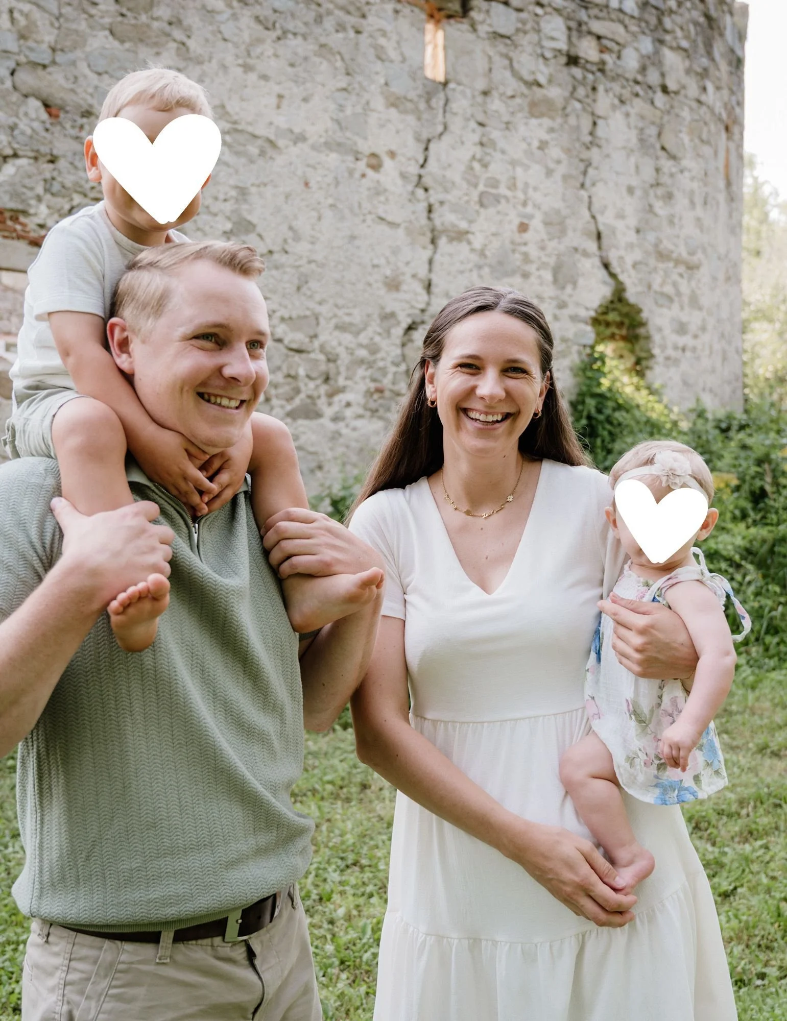 Eine lachende Familie bestehend aus einem Mann, einer Frau, einem Jungen auf den Schultern und einem kleinen Mädchen in den Armen, vor einer alten Steinmauer im Freien.