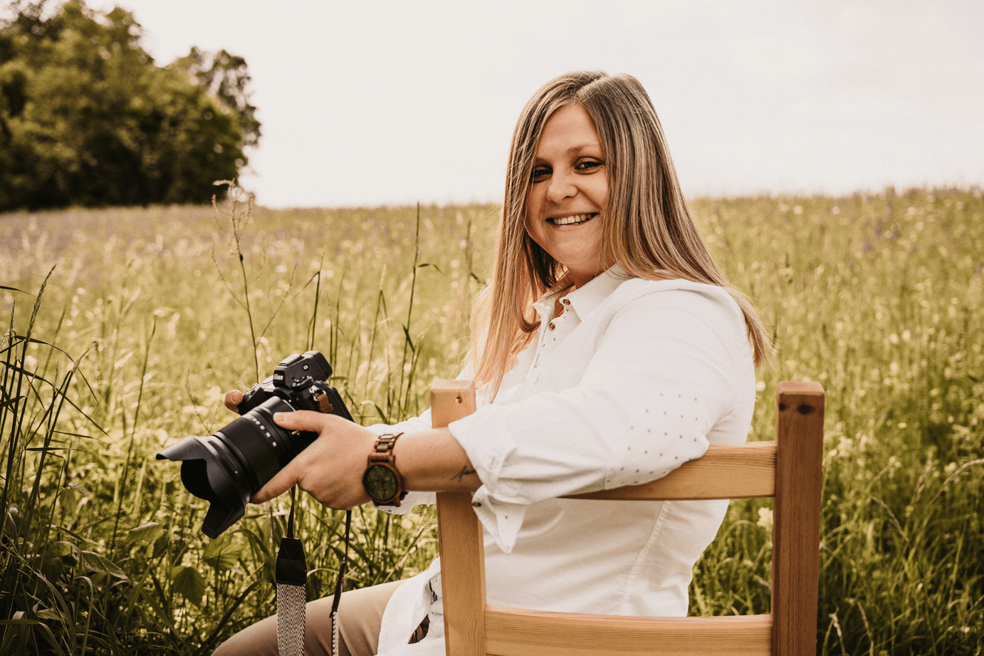 Eine lächelnde junge Frau sitzt im Freien in einem hohen Grasfeld, hält eine Kamera, trägt eine weiße Bluse, eine Armbanduhr und hat braune Haare.