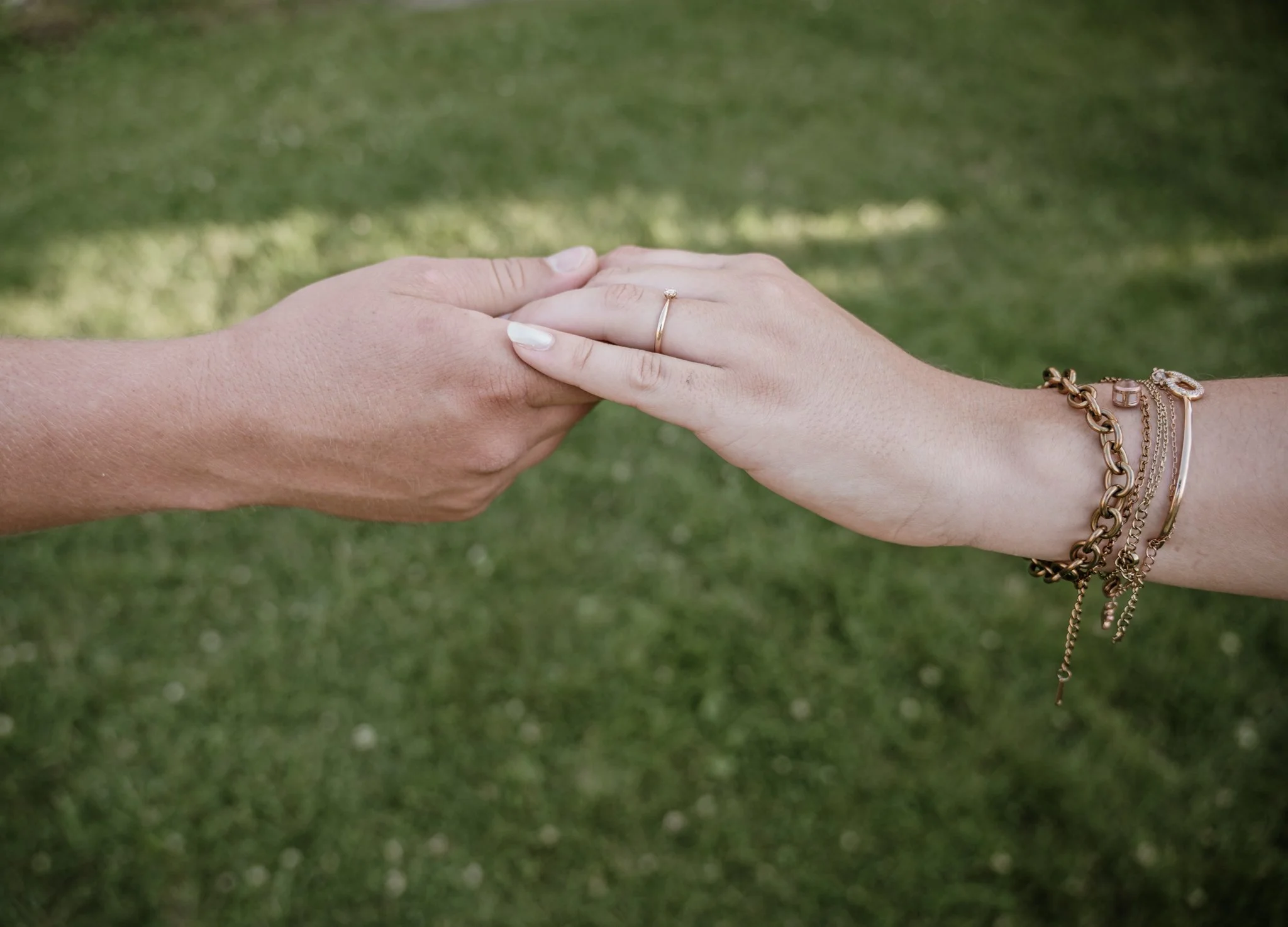 Zwei Hände halten sich an den Fingern, eine trägt einen Goldring, die andere mehrere Armbänder, auf grünem Gras im Hintergrund.
