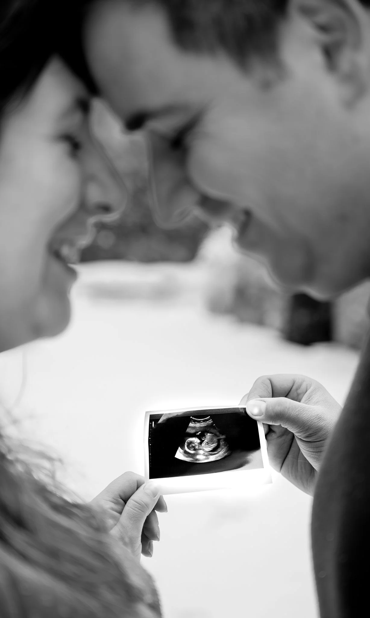 Zwei Menschen, ein Mann und eine Frau, schauen sich ein Ultraschallbild auf einem Fotobild an, wahrscheinlich ein Baby. Die Szene ist in Schwarz-Weiß gehalten.