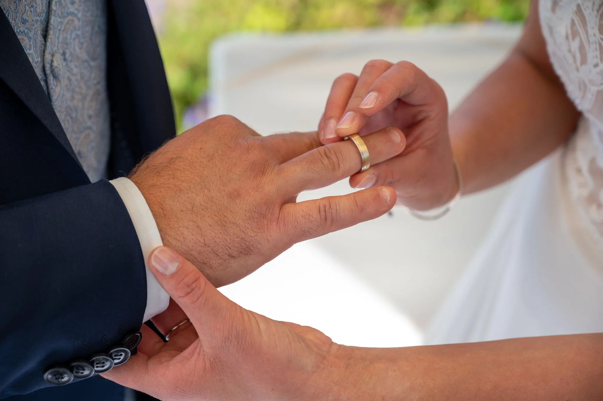 Ein Mann in einem dunklen Anzug mit weißen Manschetten legt seiner Partnerin einen Ehering an den Finger, bei ihrer Hochzeitszeremonie.
