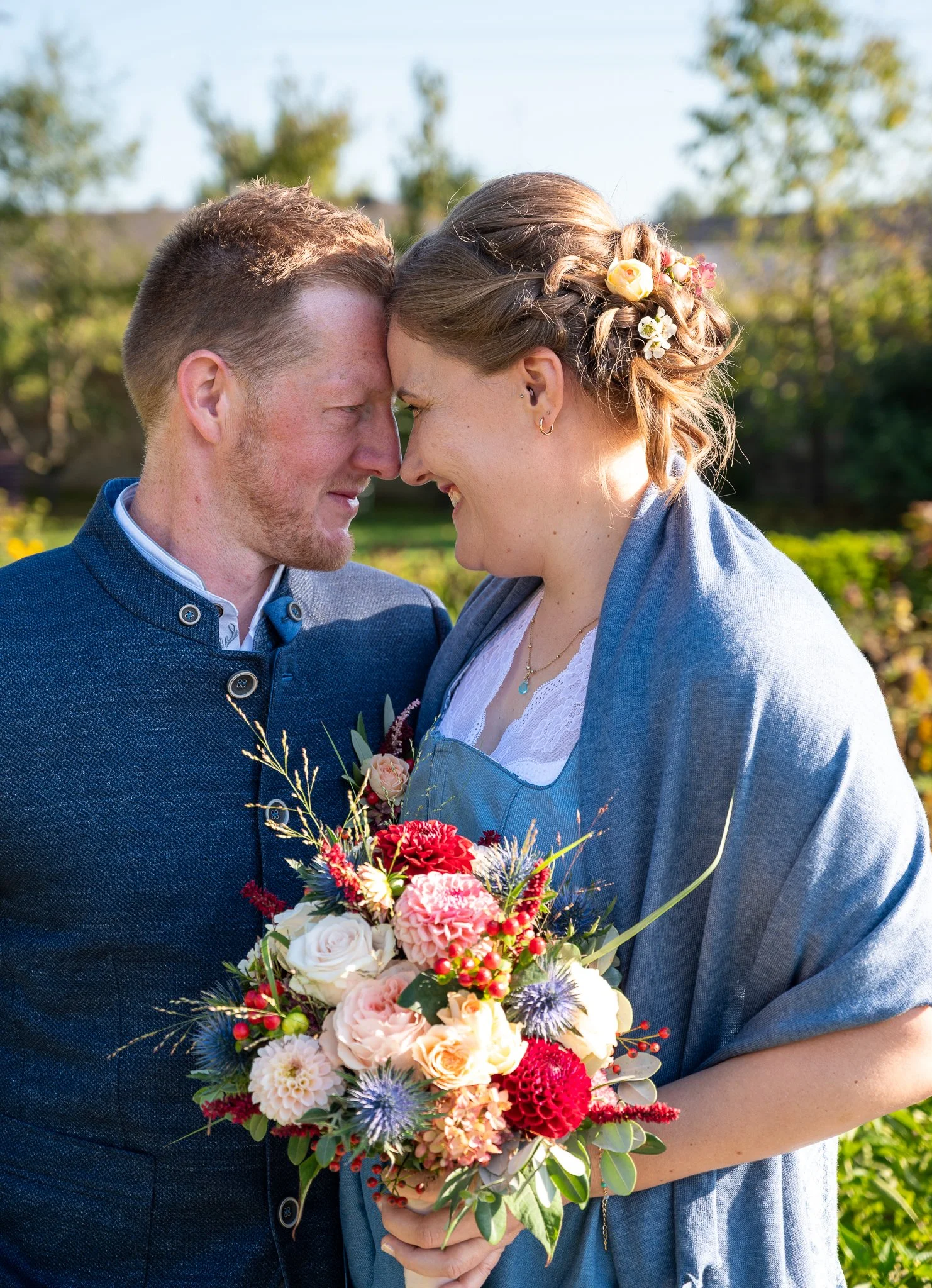 Ein Paar steht sich im Freien gegenüber, lächelnd und mit der Stirn aneinander, während die Frau einen Blumenstrauß hält.