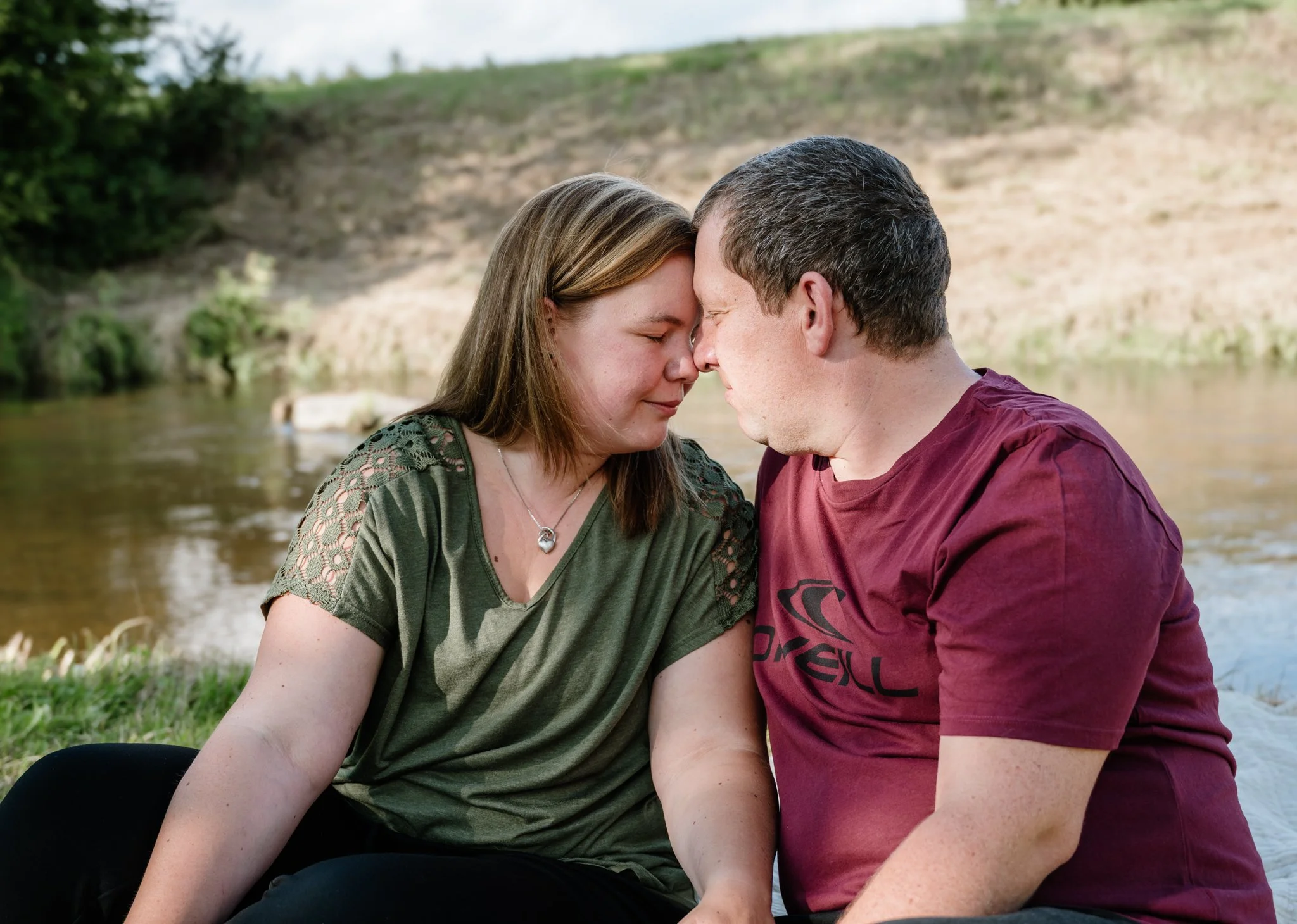 Ein Paar sitzt eng aneinander an einem Flussufer, ihre Stirne berühren sich, während sie einen intimen Moment teilen.