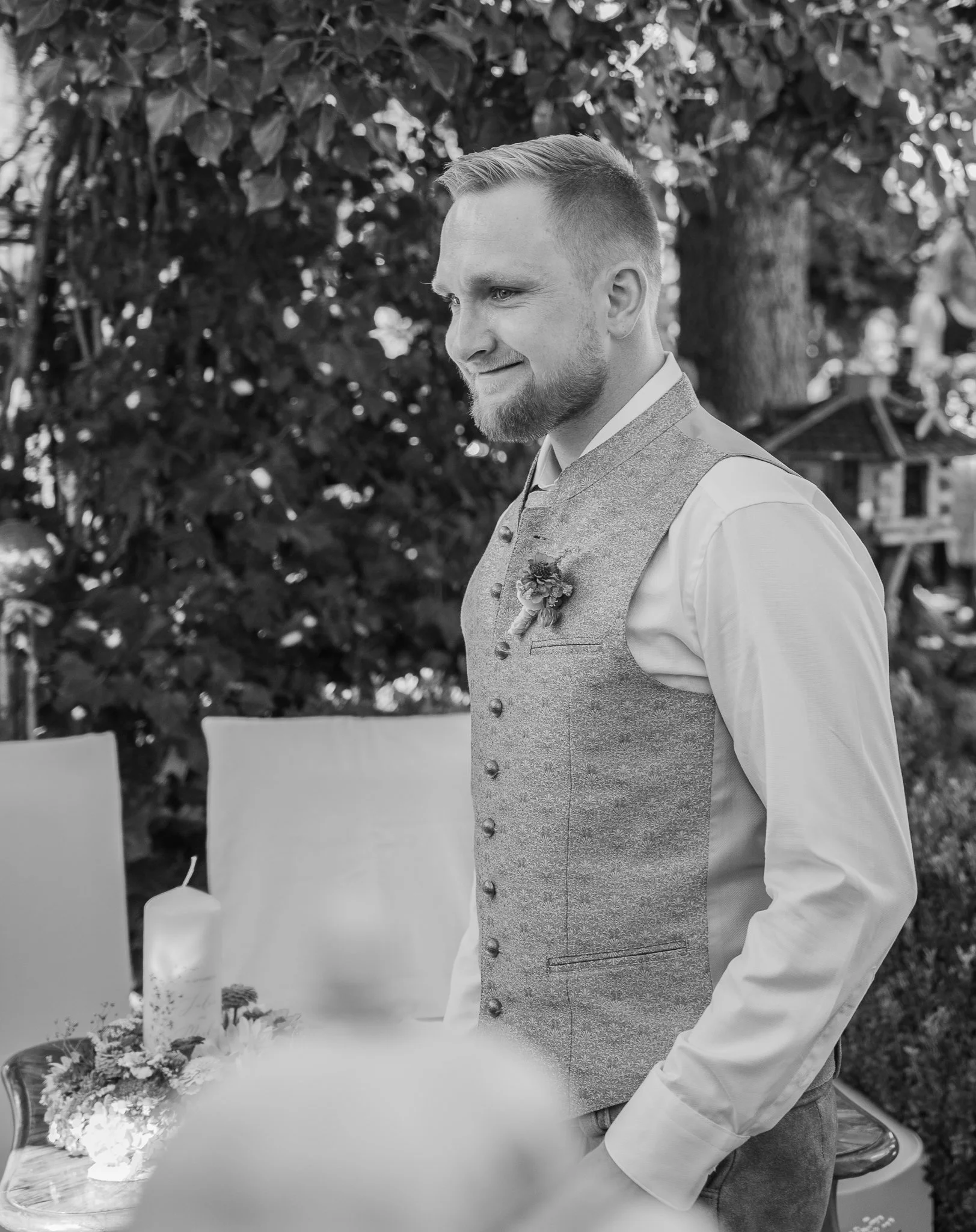 Junger Mann in formeller Kleidung bei einer Zeremonie, wahrscheinlich einer Hochzeit, im Freien mit Bäumen im Hintergrund.