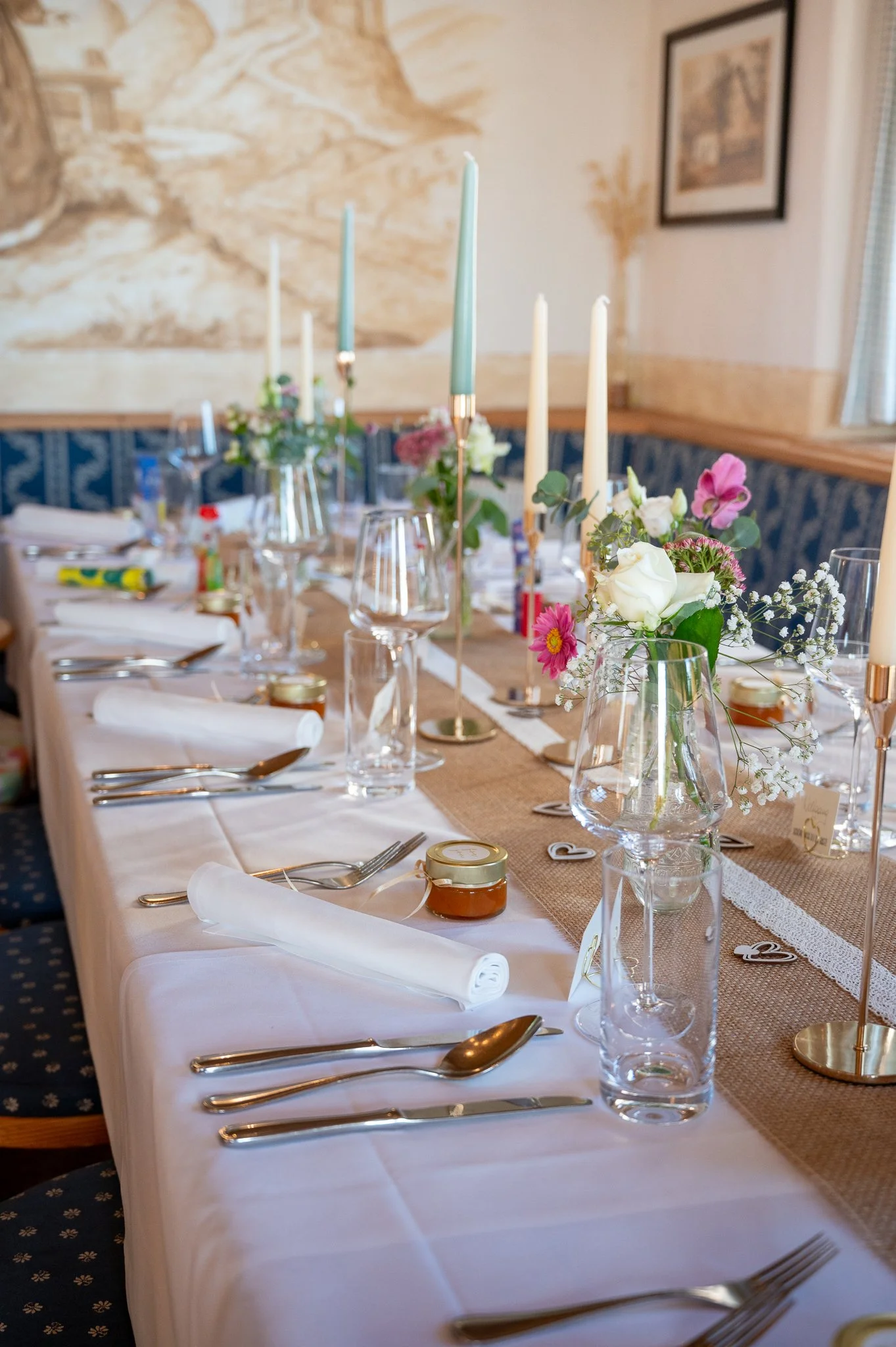 Ein festlich gedeckter Tisch mit weißen Tischdecken, Blumenarrangements in Gläsern, Kerzen, Gläsern, Besteck und kleinen Geschenken für eine Feier oder Hochzeit.
