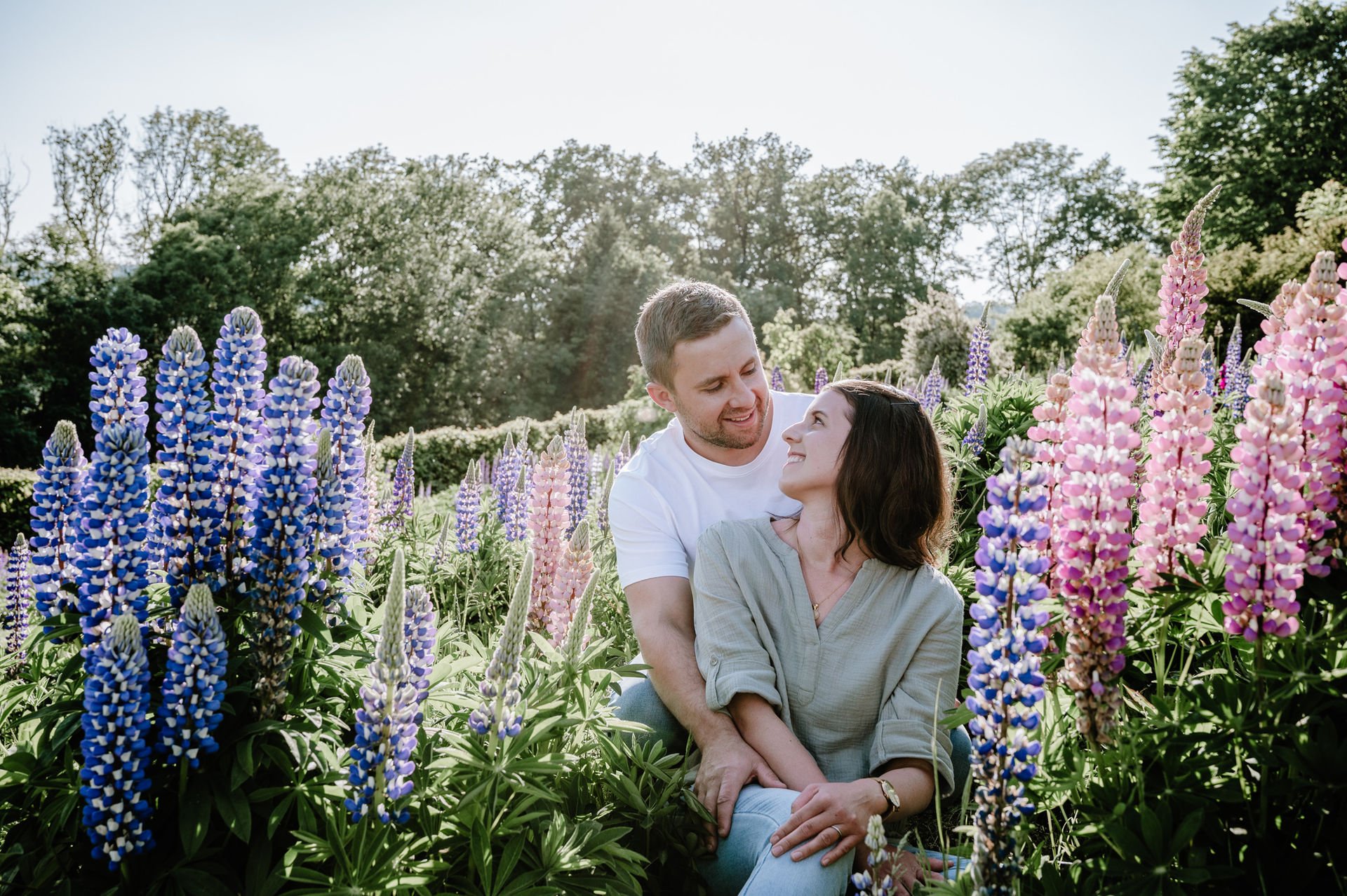 Ein Paar sitzt zwischen bunten Lupinenblumen in einem Garten, schaut sich liebevoll an, im Hintergrund bunte Bäume und Himmel.
