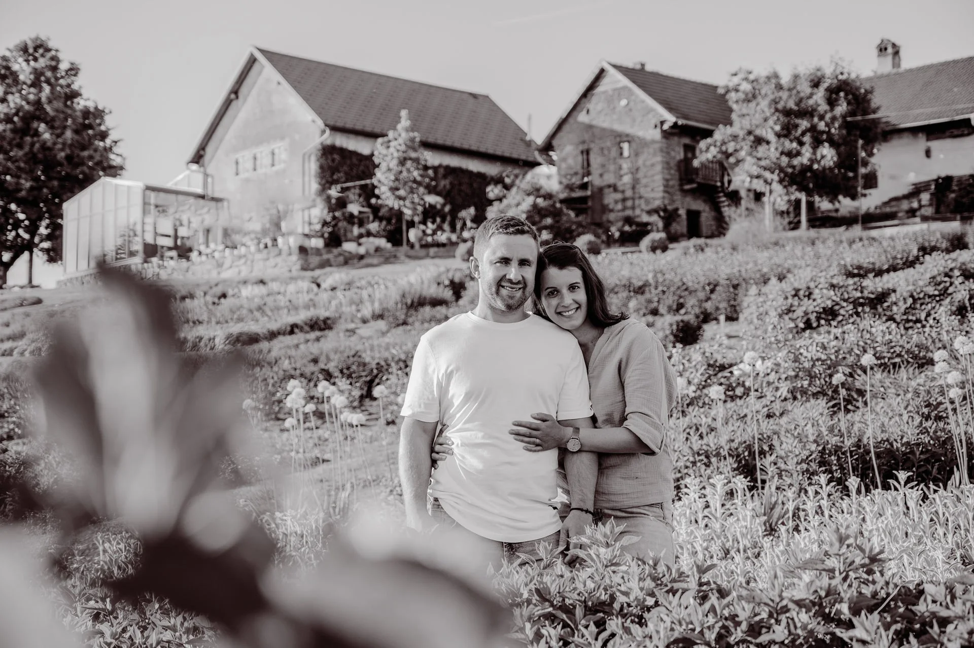 Ein glückliches Paar steht in einem Garten mit Blumen, im Hintergrund sind Häuser und Bäume. Es ist eine Schwarz-Weiß-Fotografie.