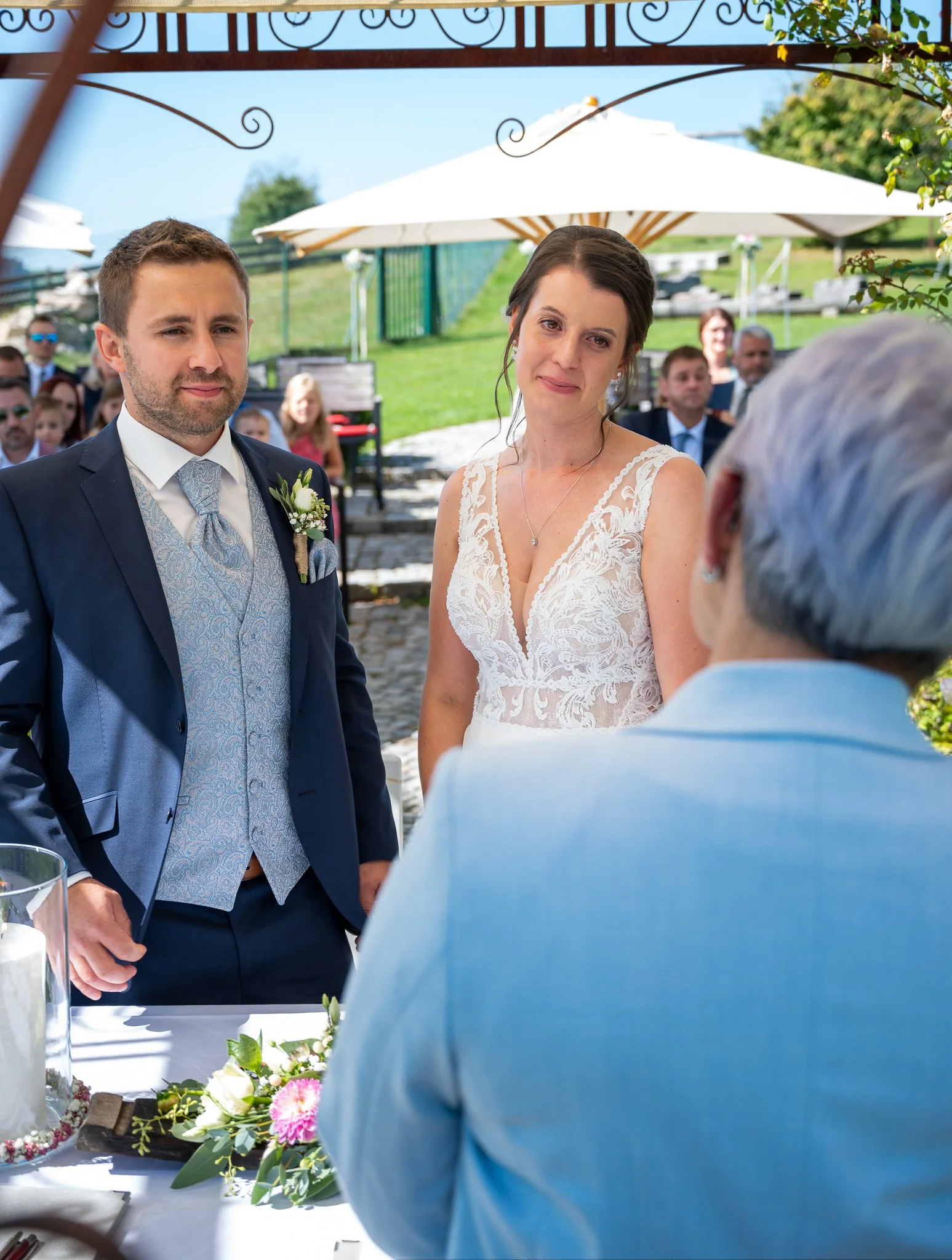 Ein Brautpaar bei einer Hochzeitszeremonie im Freien, mit Gästen im Hintergrund. Der Bräutigam trägt einen dunklen Anzug, die Braut ein weißes Kleid, und eine ältere Person steht vor ihnen, vermutlich der/die Standesbeamte oder Trauzeuge.