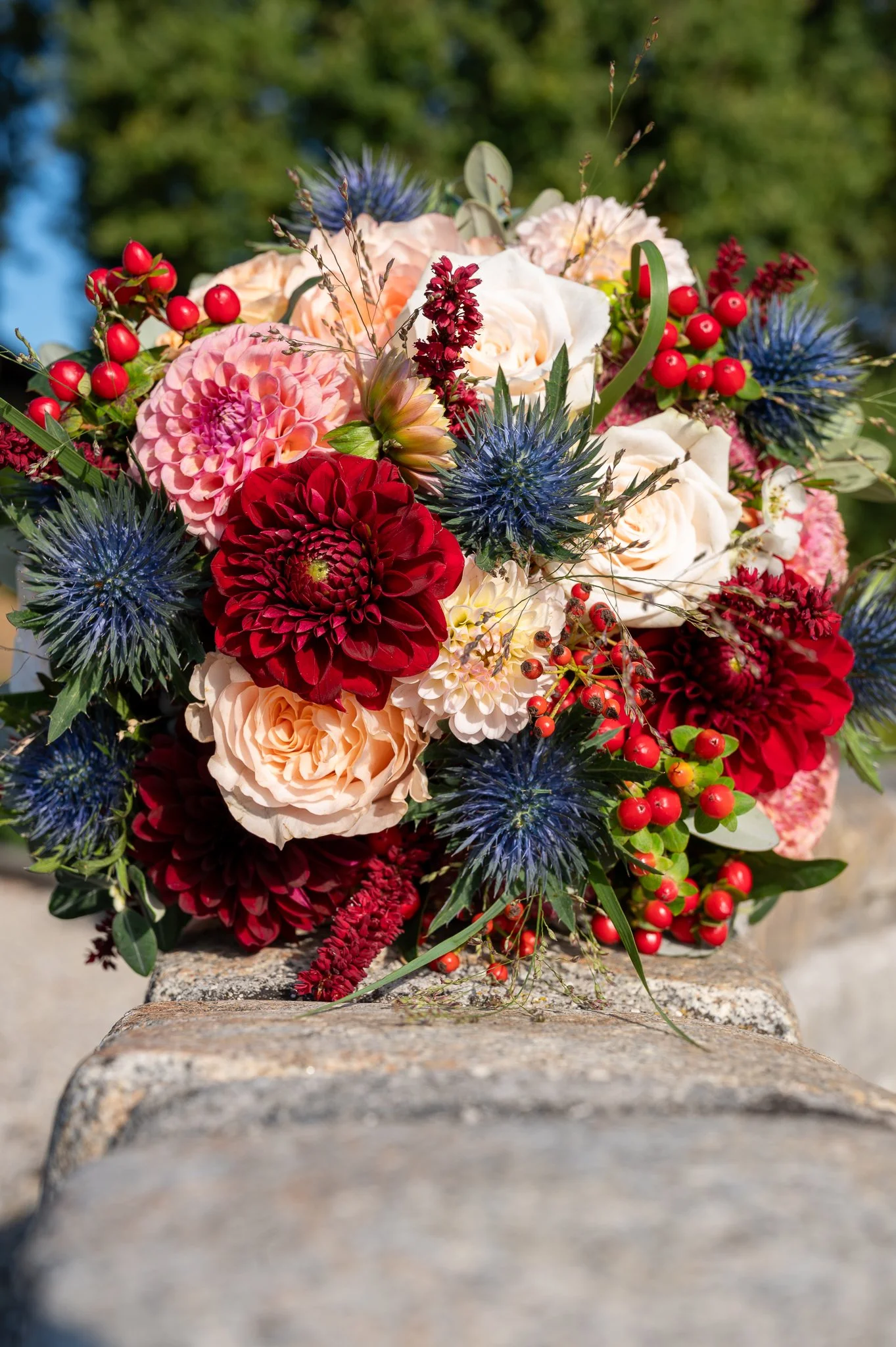 Blumengesteck mit roten, weißen und pinkfarbenen Blumen, blauen Disteln und roten Beeren auf einem Stein