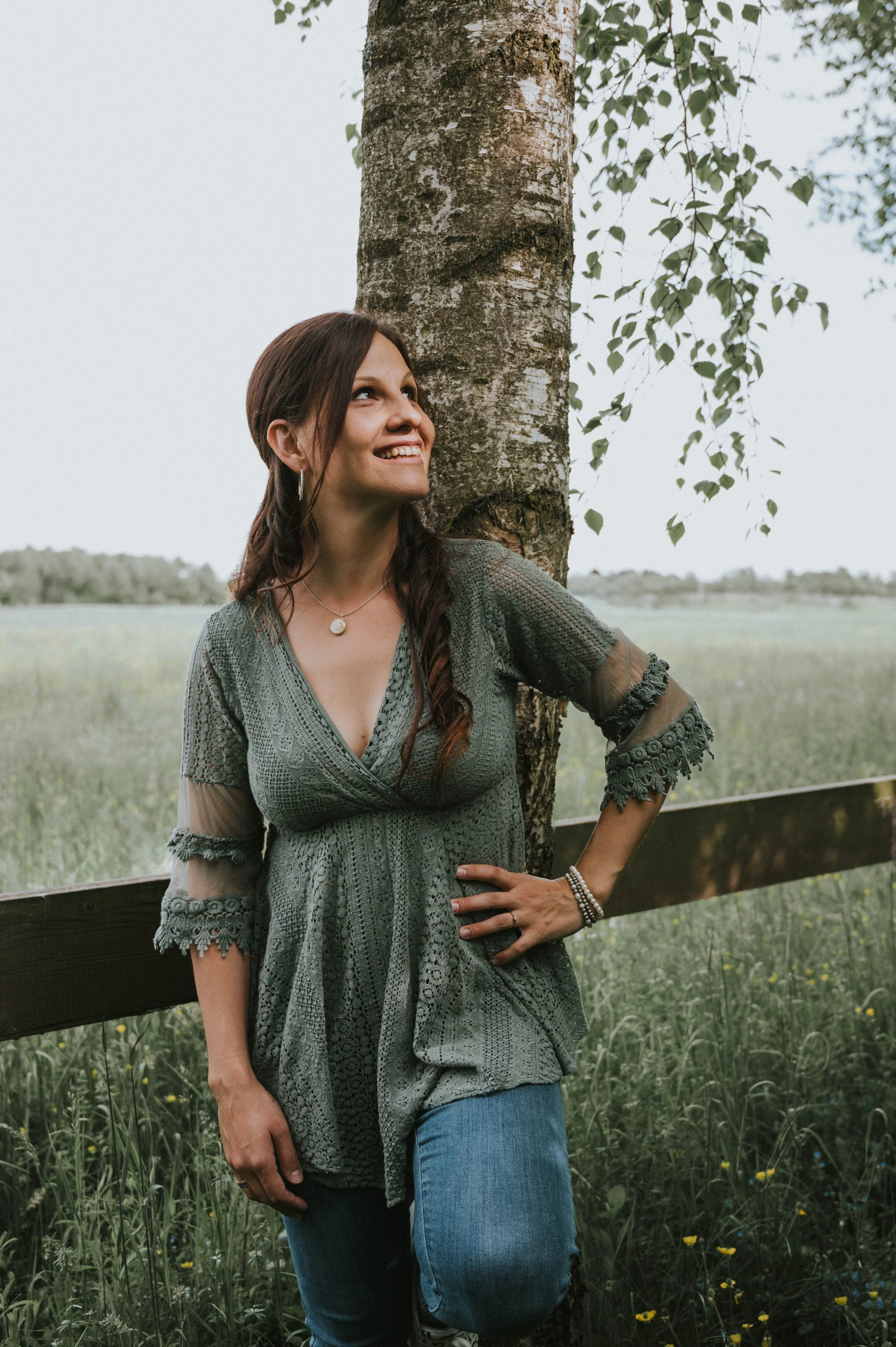 Eine Frau lehnt an einem Baum in einer grünen Wiese mit Blick in die Ferne und lächelt.