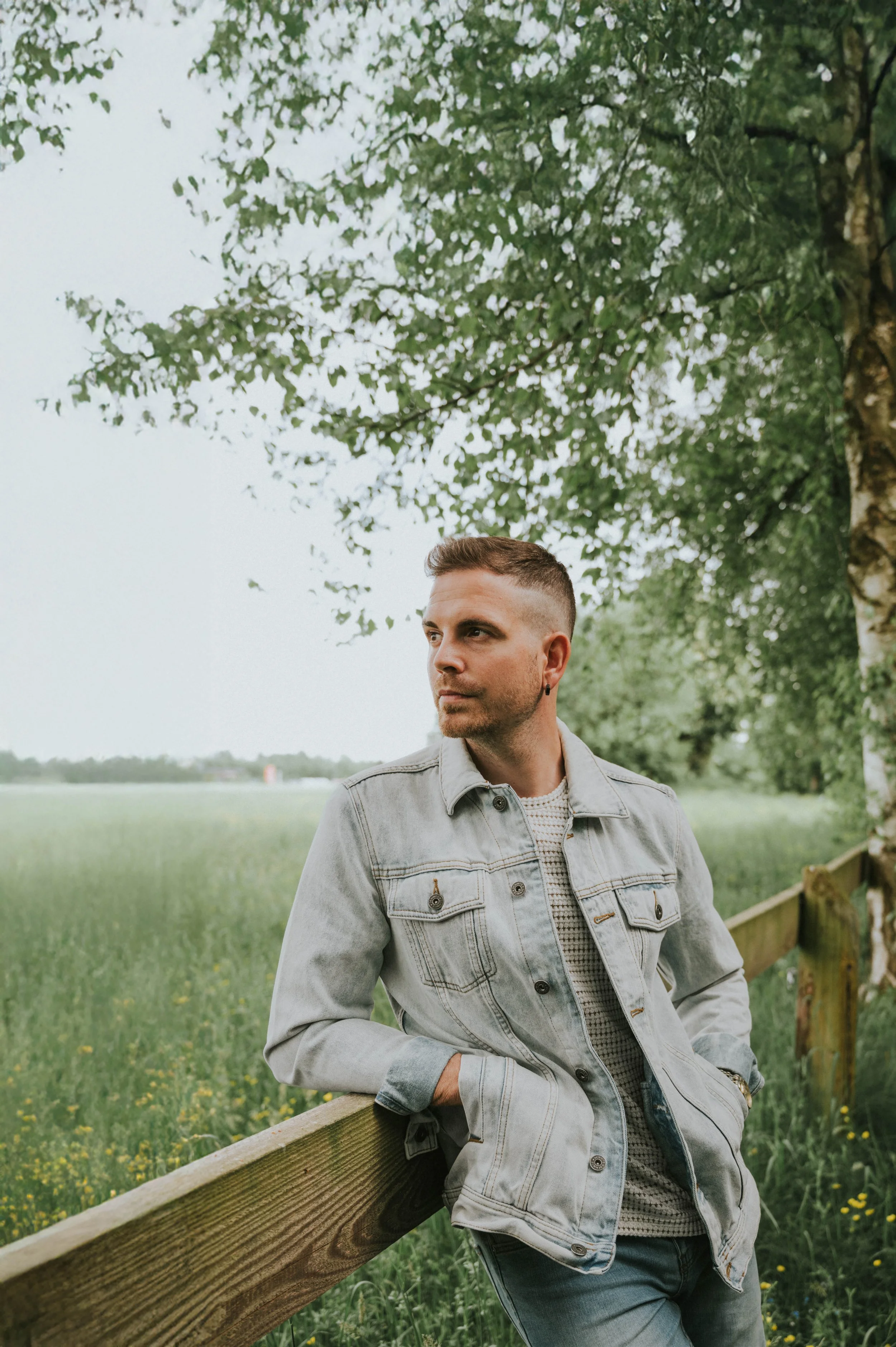 Ein junger Mann lehnt an einem Holzzaun in einer grünen Wiese, unter einem Baum, bei bewölktem Himmel.