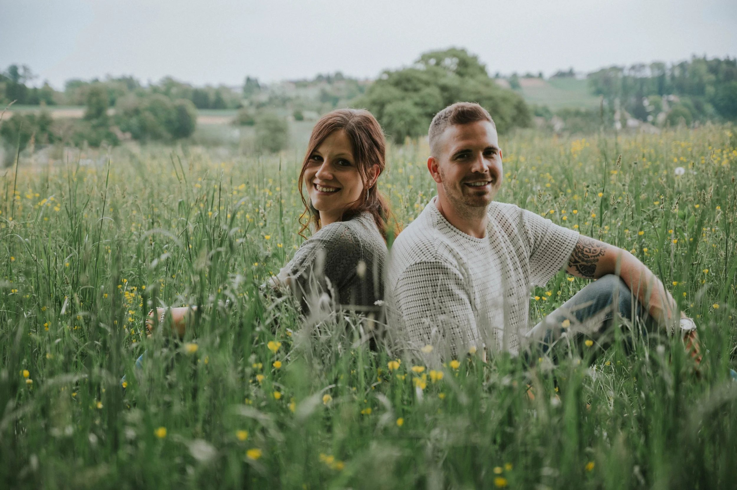 Ein lachendes Paar sitzt in einem grünen Feld mit gelben Blumen, Rücken an Rücken, bei sonnigem Wetter.