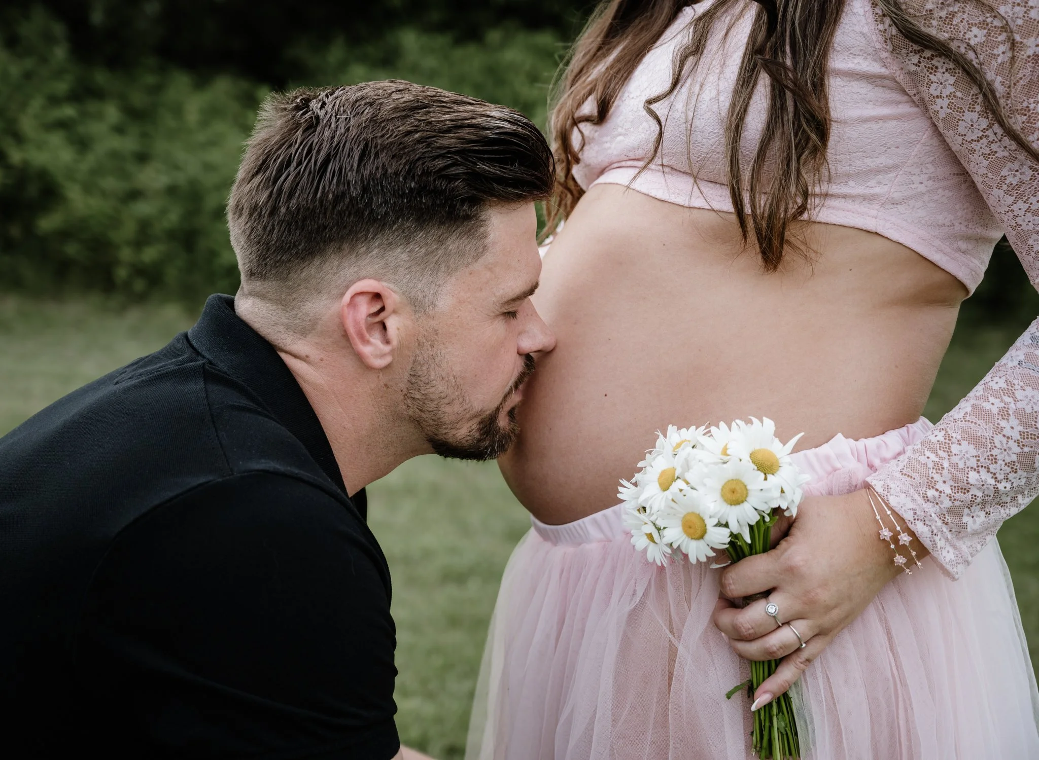 Ein Mann küsst eine schwangere Frau auf den Bauch während eines Fotoshootings im Freien, die Frau hält einen Blumenstrauß aus Gänseblümchen.