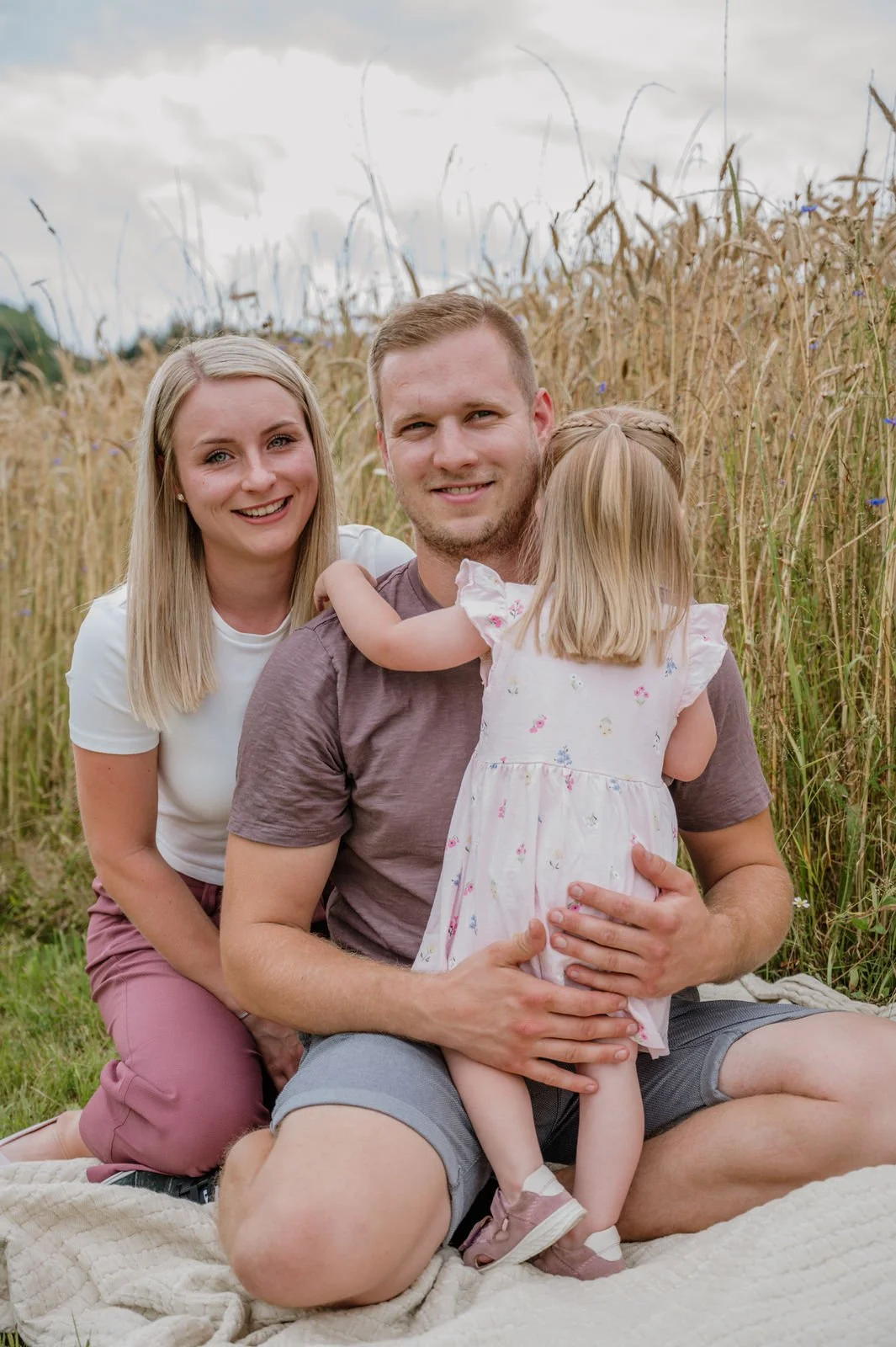 Familie sitzt in einem Getreidefeld, Mutter, Vater und kleines Mädchen, lachend und umarmend.