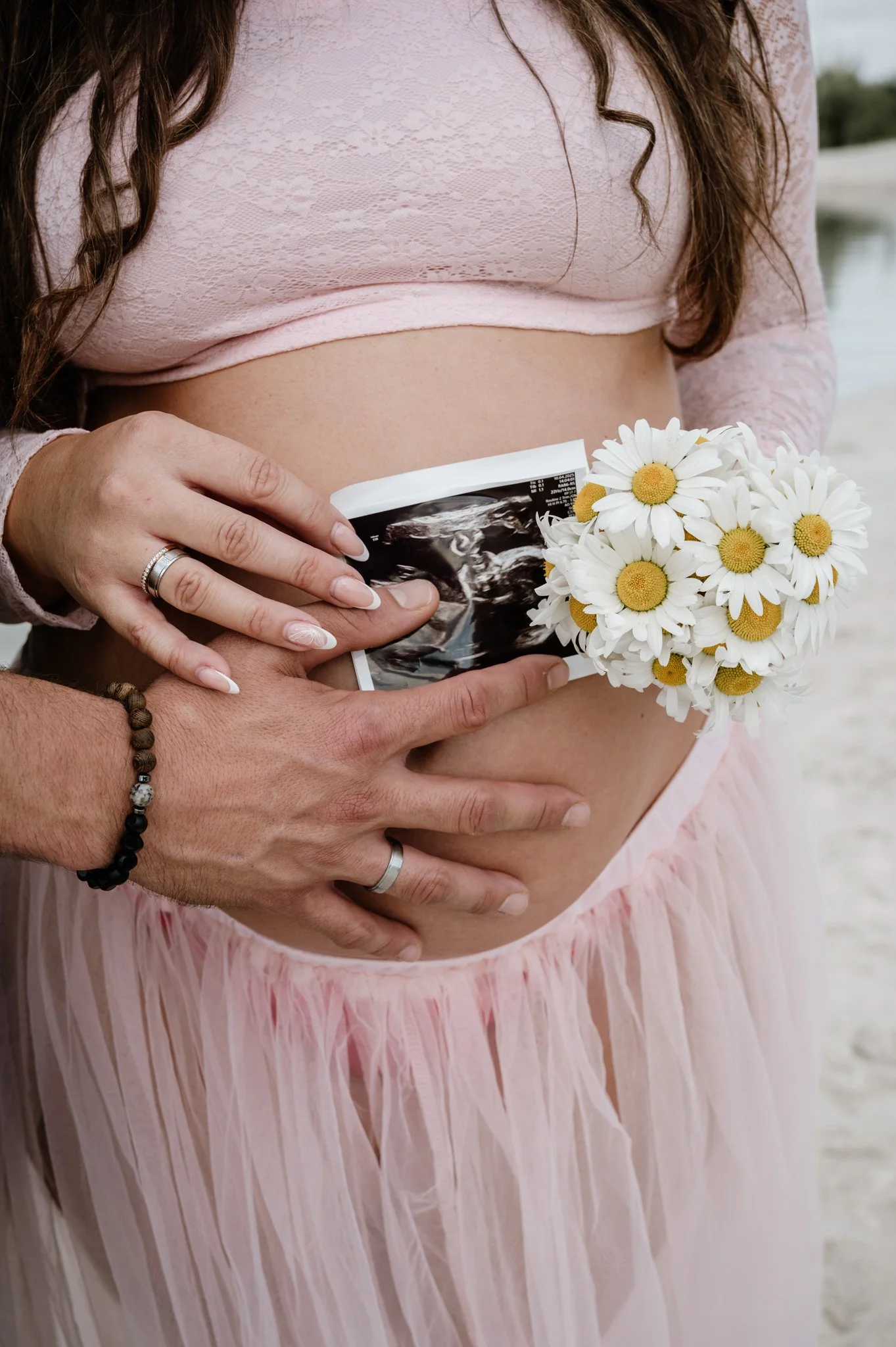 Schwangere Frau hält ein Ultraschallbild und einen Blumenstrauß mit Gänseblümchen, während eine Hand von einem Partner die Bauchregion berührt.