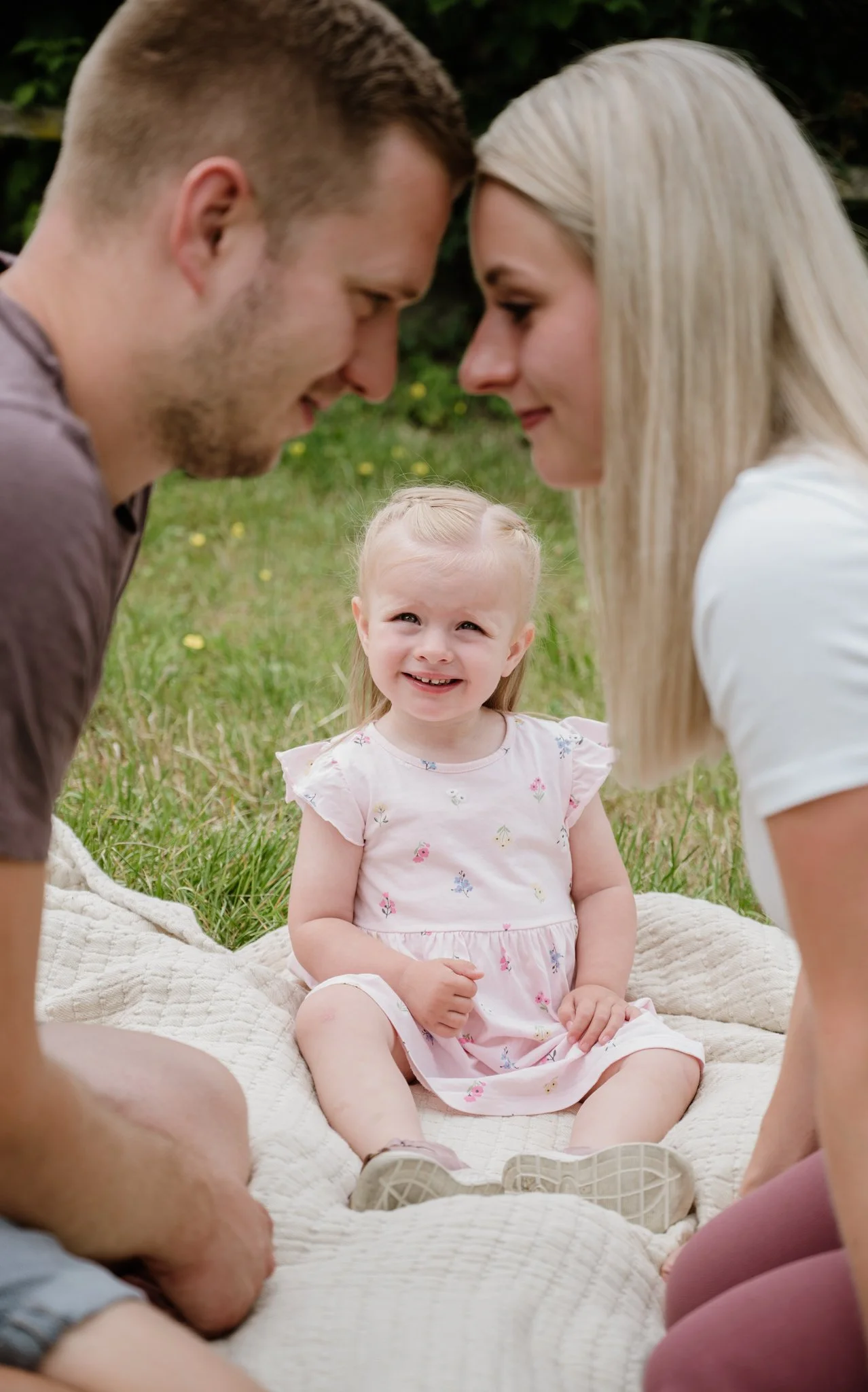 Ein junges Mädchen sitzt auf einer Decke im Freien, umgeben von einem Mann und einer Frau, die sich mit ihr berühren und ihr in die Augen schauen, während sie auf einer Wiese mit Bäumen im Hintergrund sitzen. Das Mädchen trägt ein rosa Kleid mit Blum